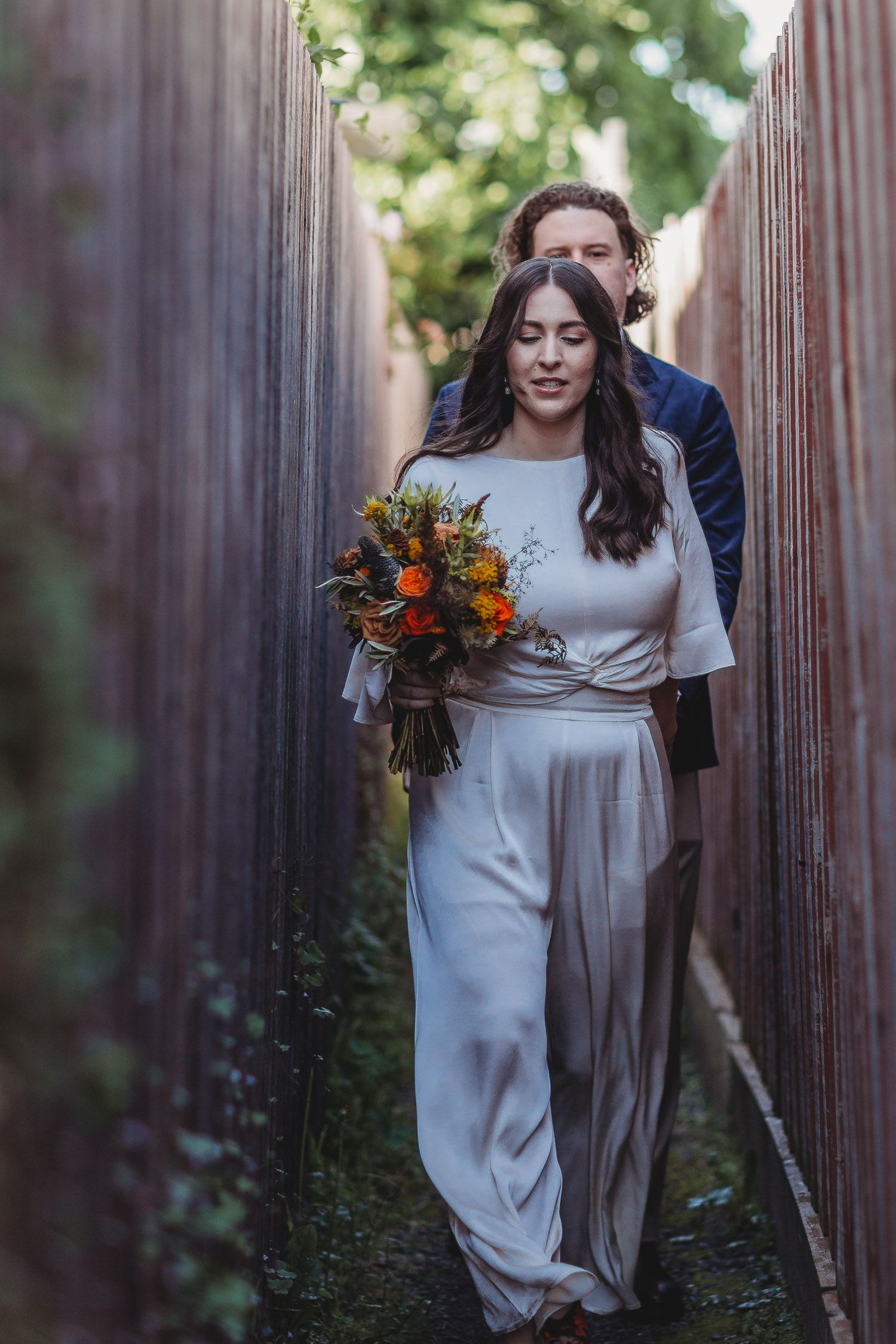 A bride in a flowing white satin outfit holds an orange autumn bouquet as she walks down a narrow wooden fence path with a man behind her.