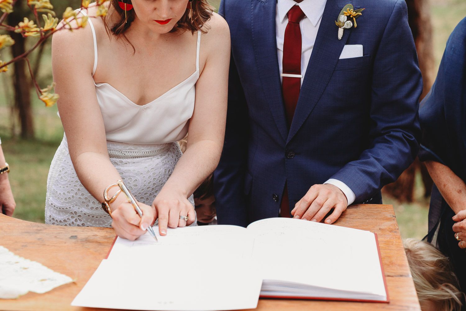 Bride signing wedding register at outdoor ceremony beside groom in blue suit.