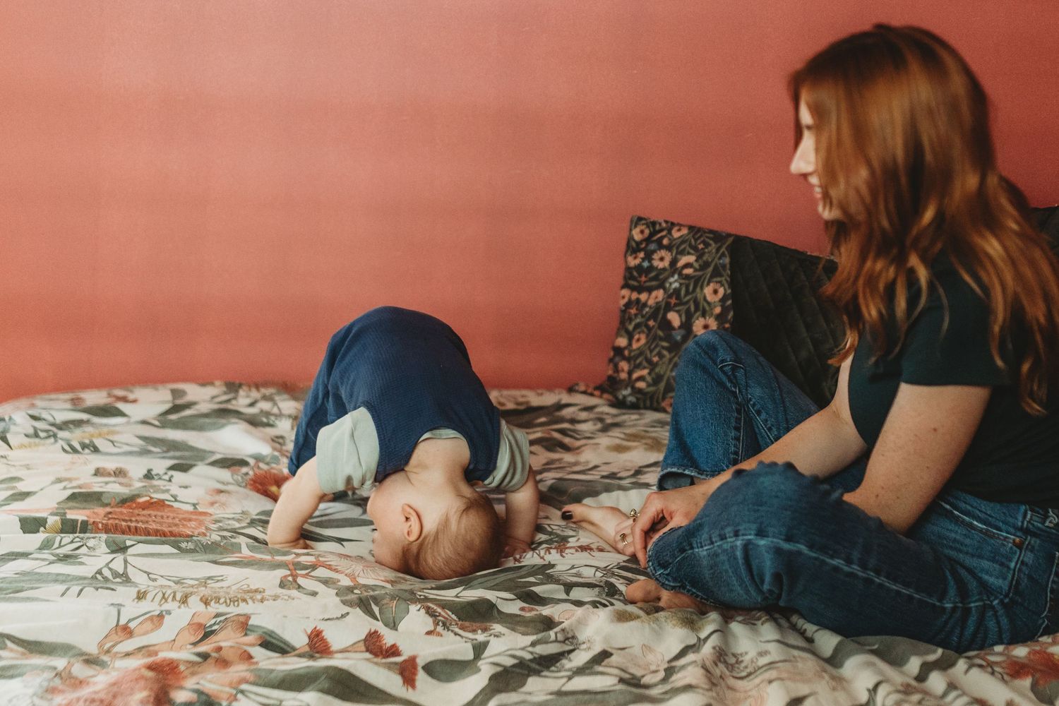 A red-haired woman in jeans sits on a floral bedspread beside a toddler doing a playful headstand against a coral pink wall.