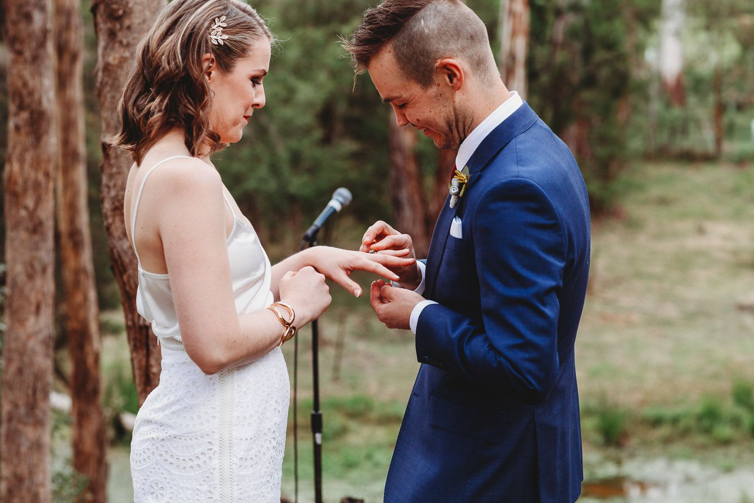 Groom placing wedding ring on brides finger during outdoor forest ceremony.