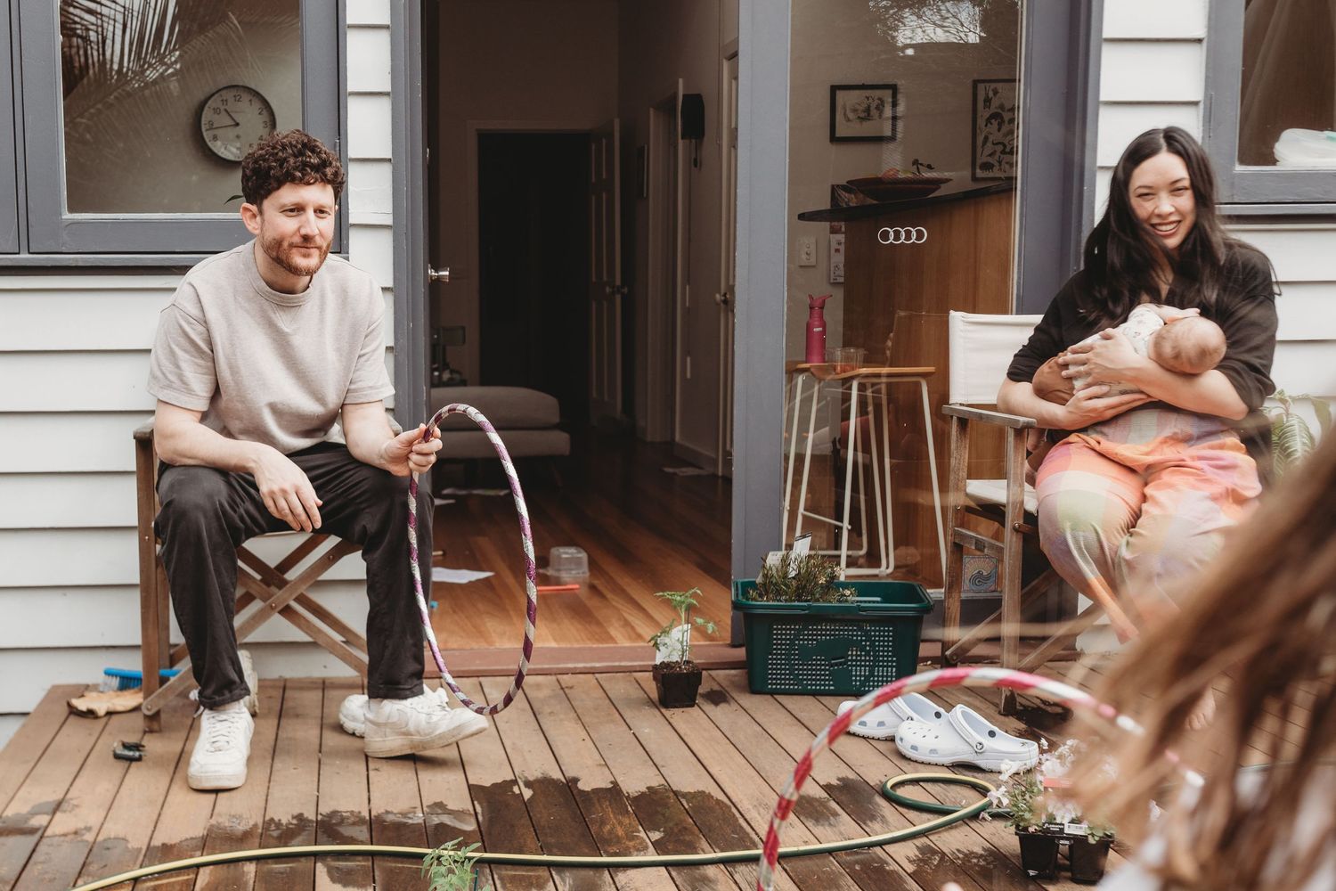 A smiling man sits on a wooden deck holding a hose while a woman cradles a baby nearby, with potted plants and a cozy house behind them.