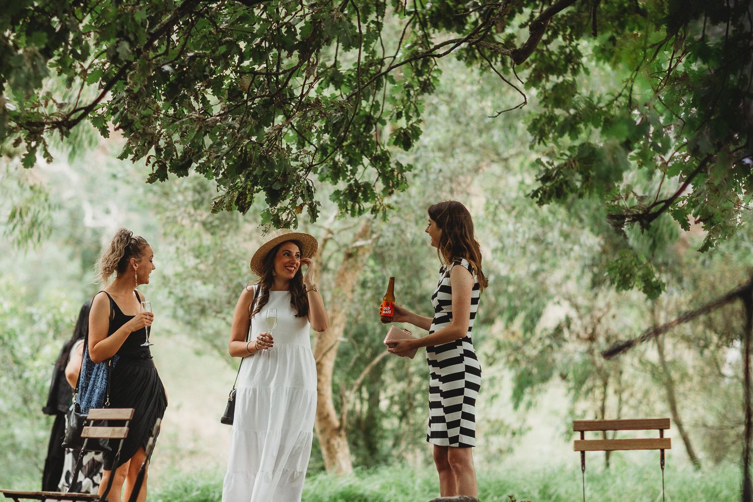 Three women in summer dresses socializing outdoors under lush green trees at a wedding.