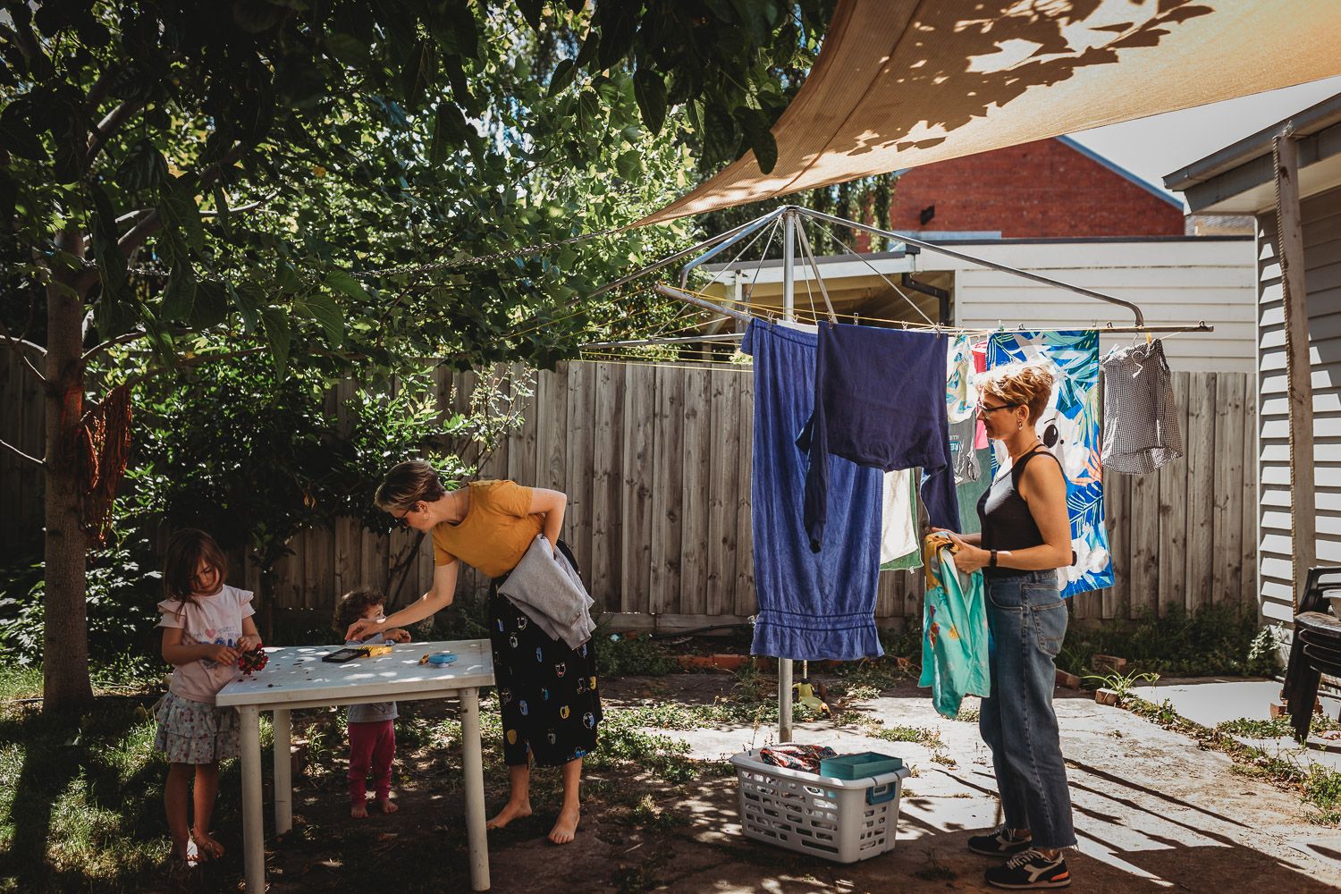 Family doing laundry outdoors in a sunny backyard with a clothesline and wooden fence.