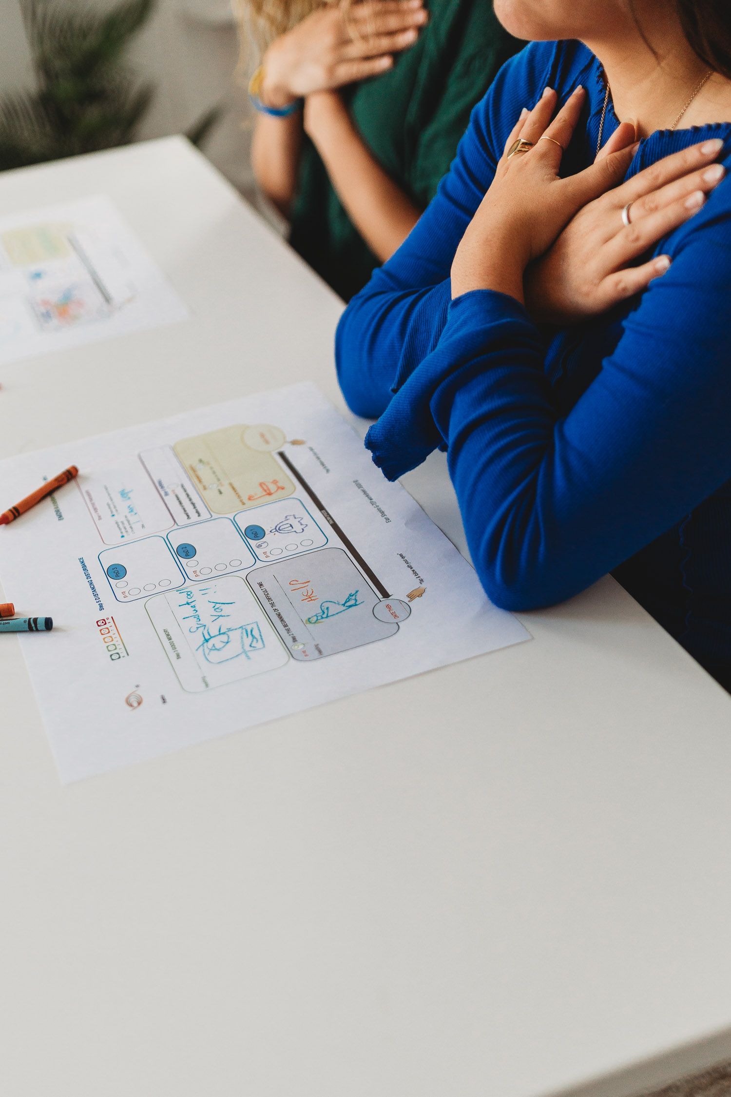 Woman in blue sweater reviewing hand-drawn UX wireframe sketches on white table with crayons.