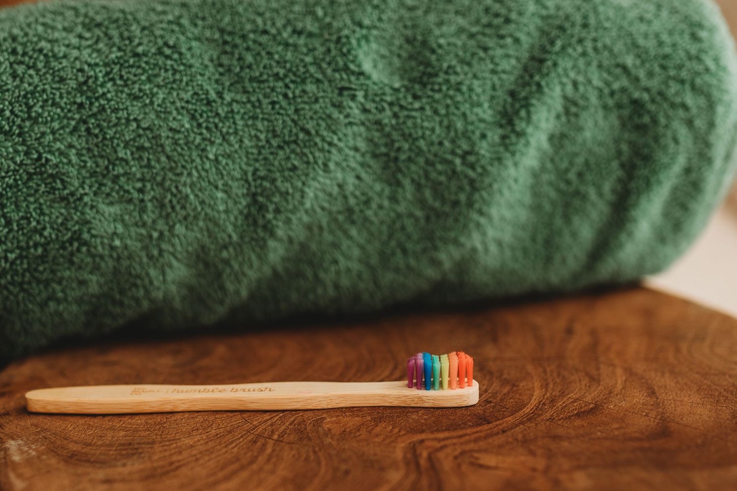 Bamboo toothbrush with rainbow bristles resting against a rolled green towel on wood surface.