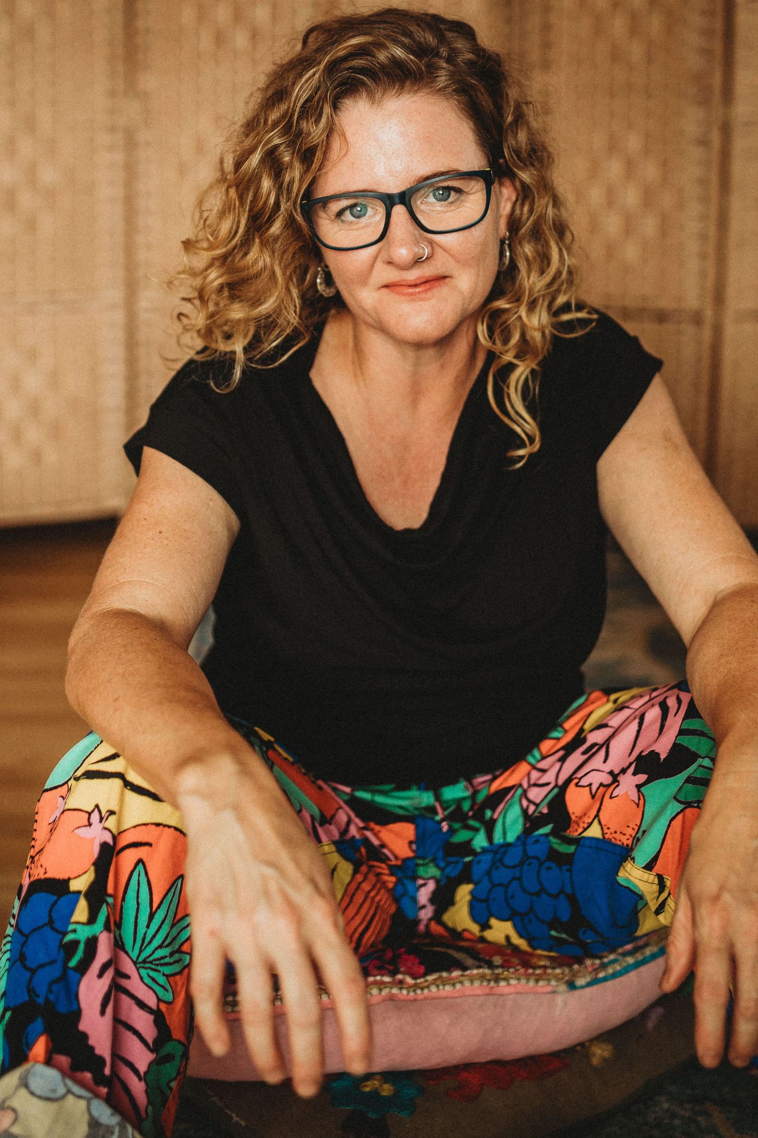 A woman with curly hair and glasses sits relaxed wearing colorful floral pants indoors.