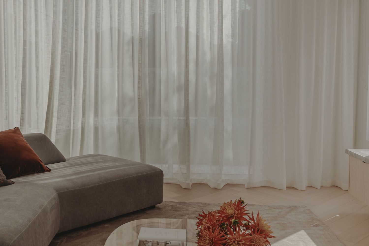 Minimalist living room with sheer white curtains, grey sofa, and terracotta cushion.