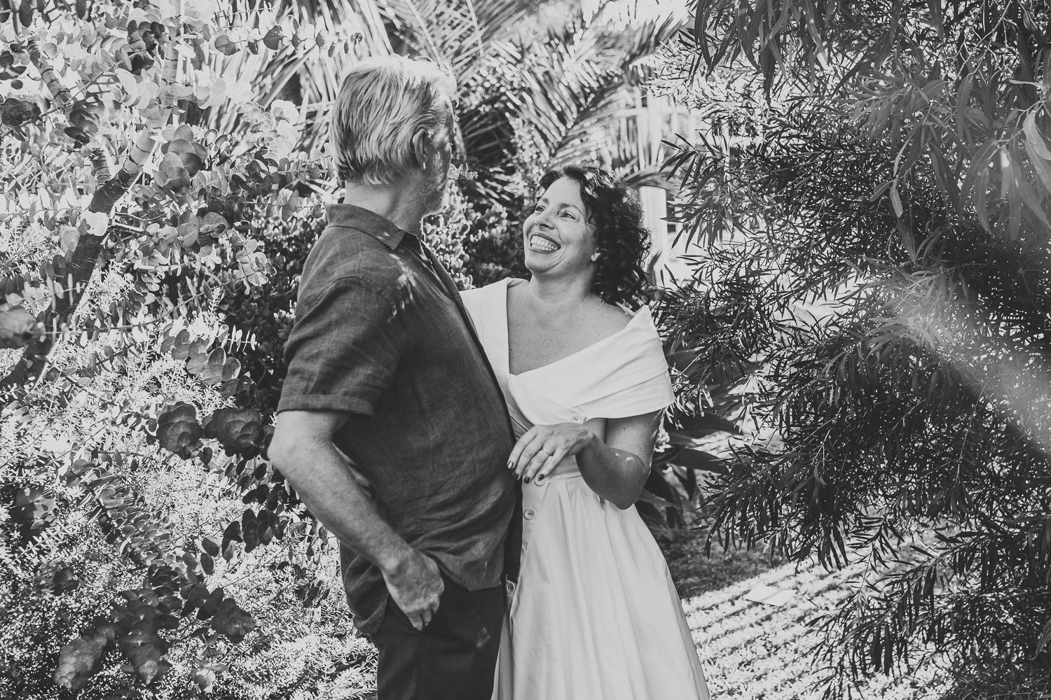 Black and white photo of a couple laughing together among tropical plants during their wedding.