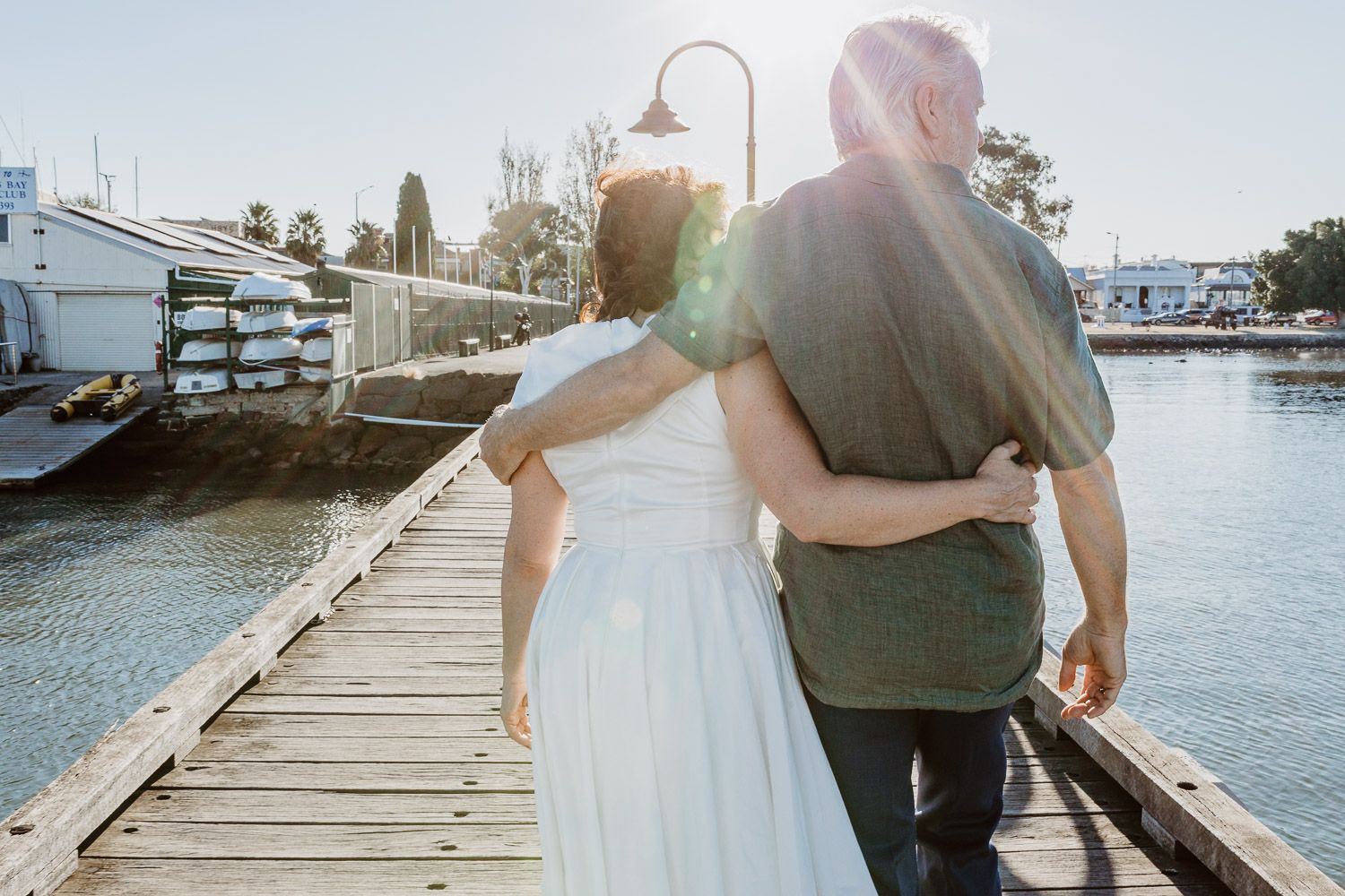 Bride and groom strolling arm in arm along a sunlit marina dock with boats in the background.