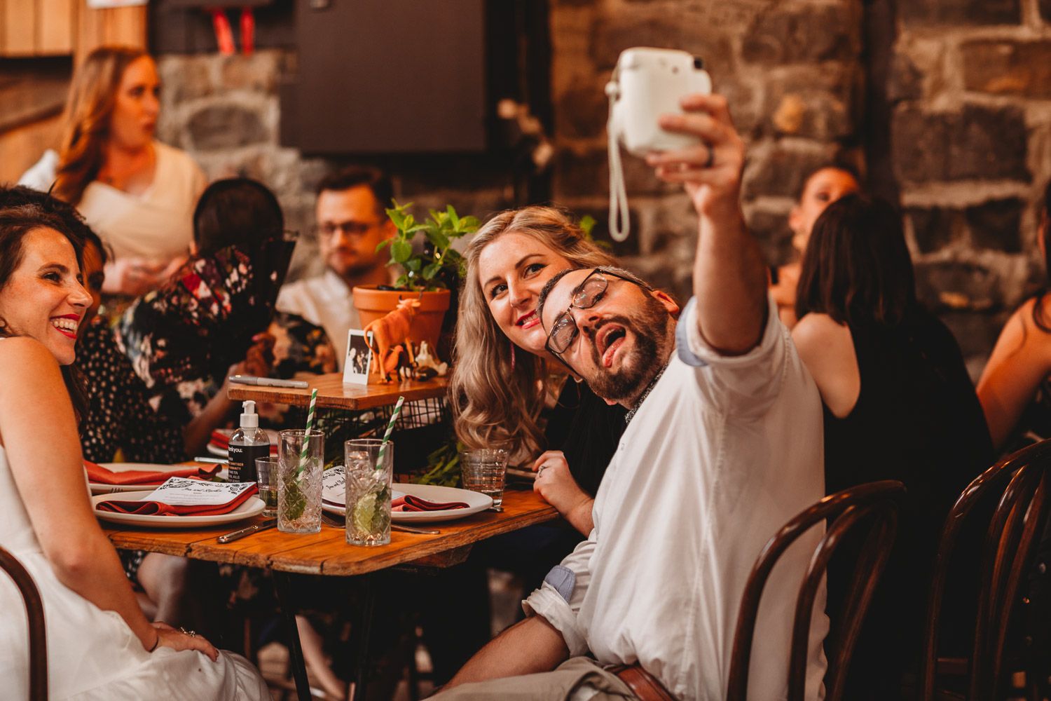 Happy couple takes a selfie with a Polaroid camera while guests laugh and celebrate at a wedding reception dinner.