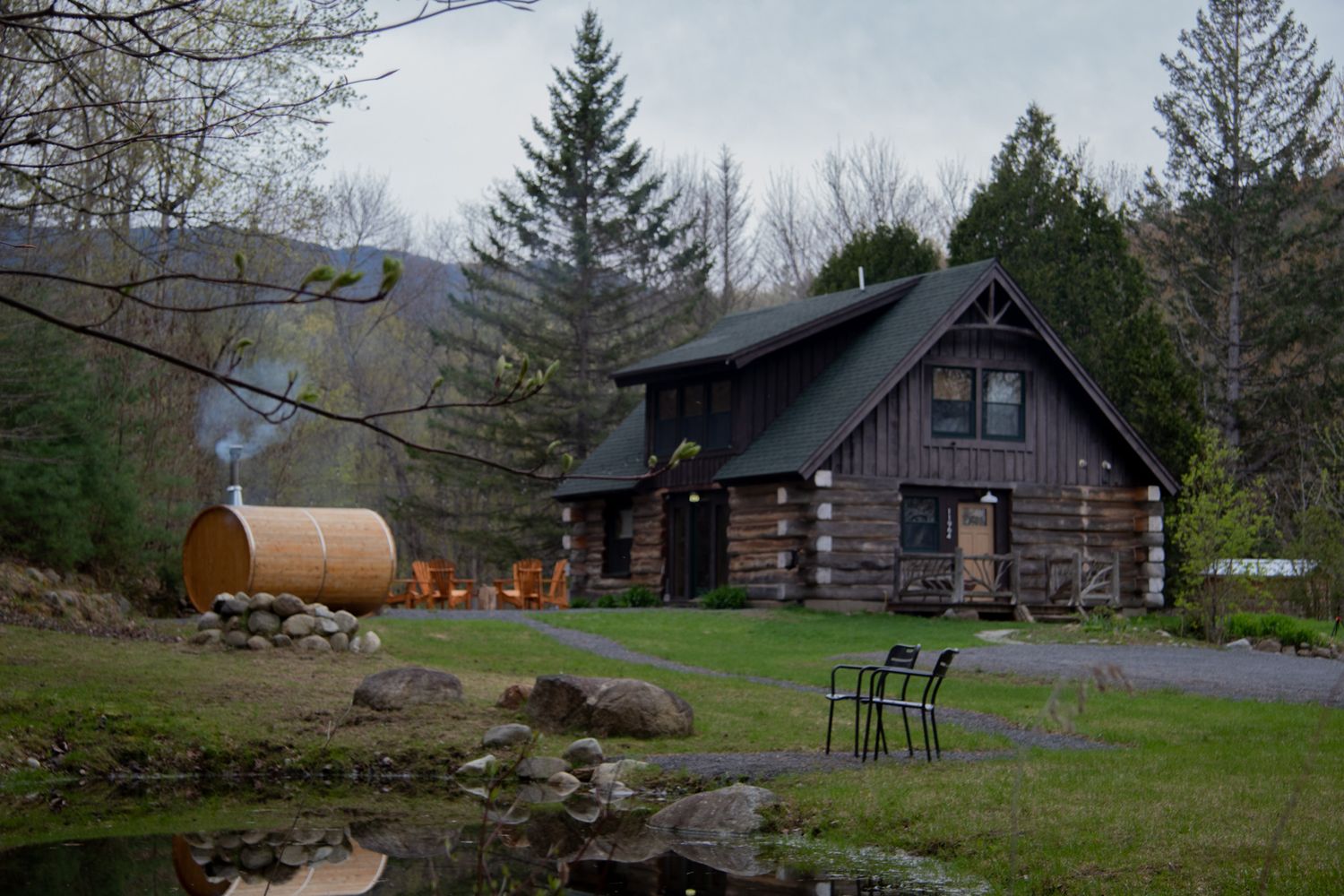 Rustic log cabin retreat with barrel sauna, Adirondack chairs, and lush green landscape surrounded by forest.