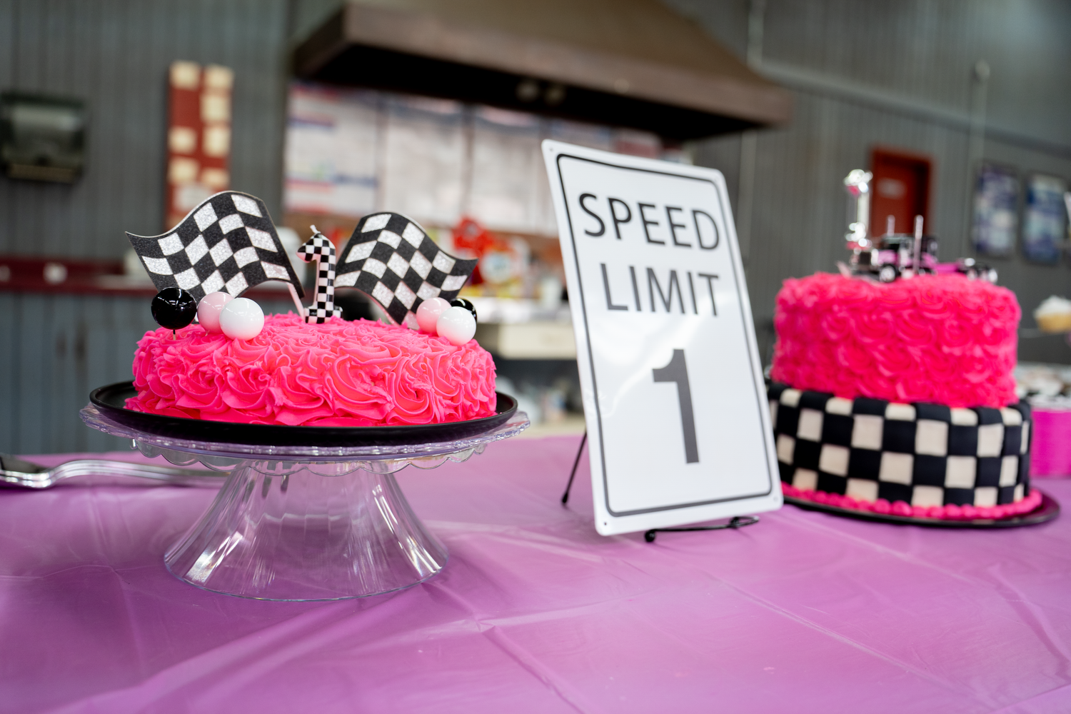 Pink rosette birthday cakes with checkered flag toppers beside a Speed Limit 1 sign on a purple tablecloth.