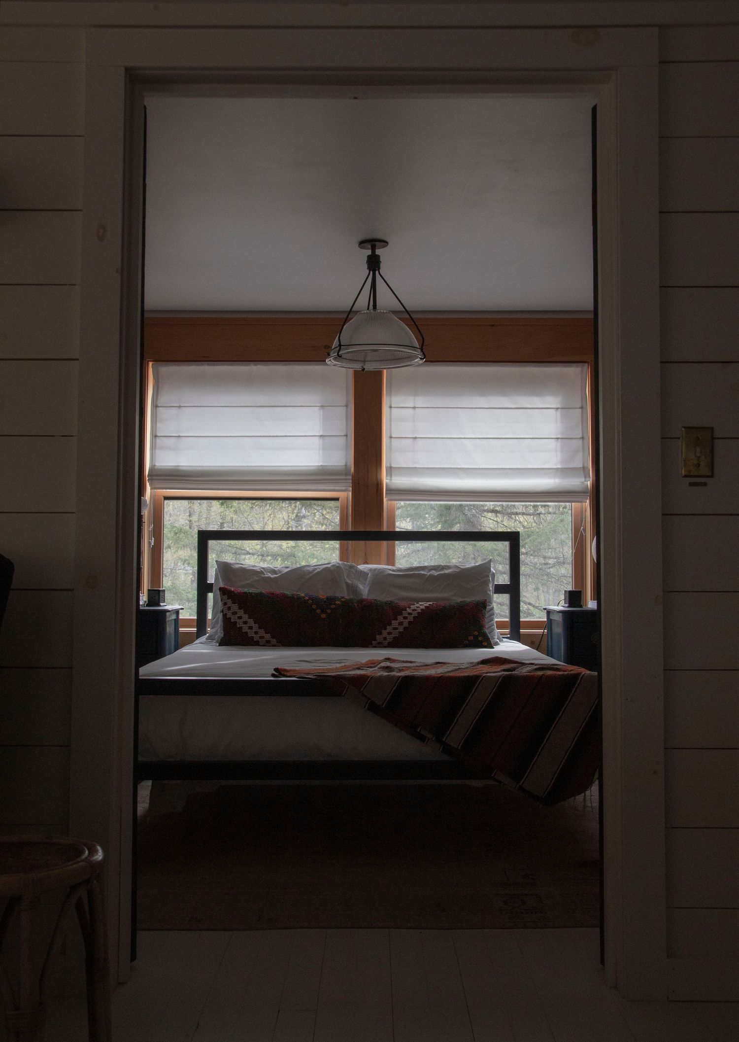 Cozy cabin bedroom viewed through doorway with metal bed frame, striped blanket, and pendant light above window.