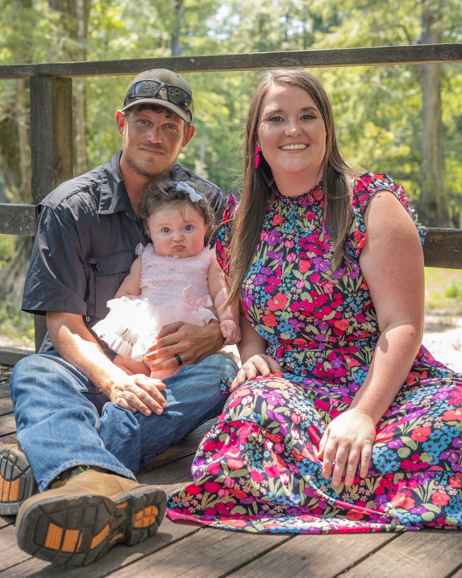 Young family with baby girl poses on wooden bridge surrounded by lush green trees.