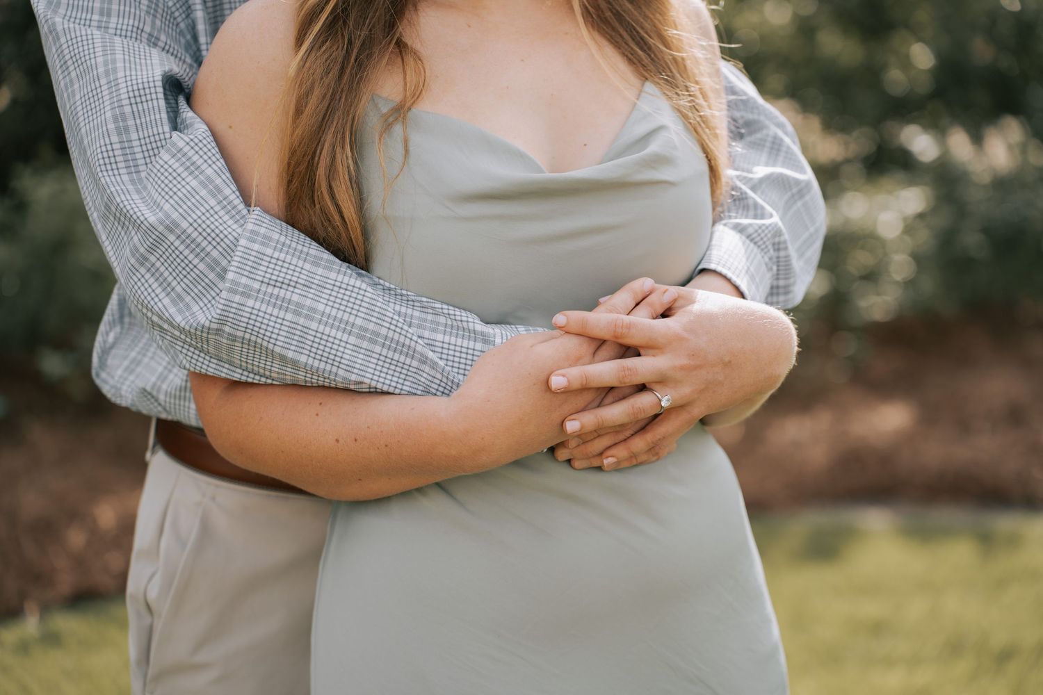 Couple embracing outdoors, man in plaid shirt wrapping arms around woman in sage green dress, hands clasped over baby bump.