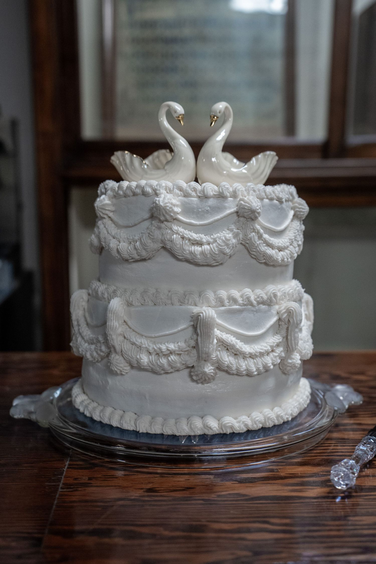 Two-tier white wedding cake with swan toppers and elegant ruffled frosting on a glass cake stand.