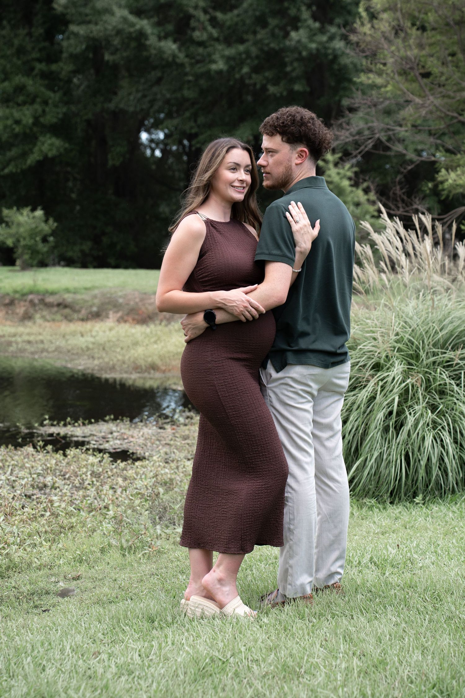 Couple sharing a tender moment outdoors near a pond, pregnant woman in brown dress smiling, man in green polo embracing her.