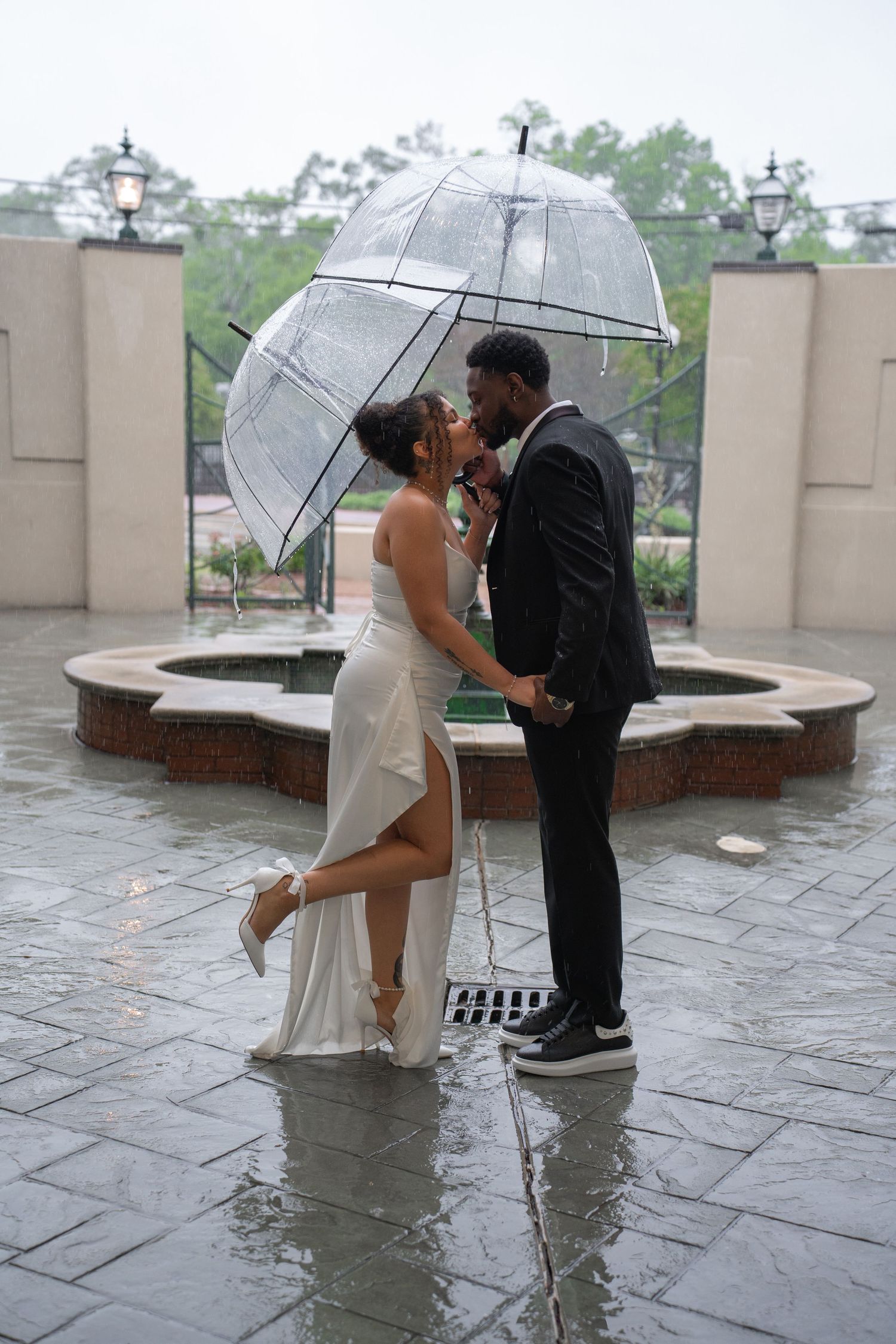 Couple kissing under clear bubble umbrella in the rain, wearing formal attire by a fountain.