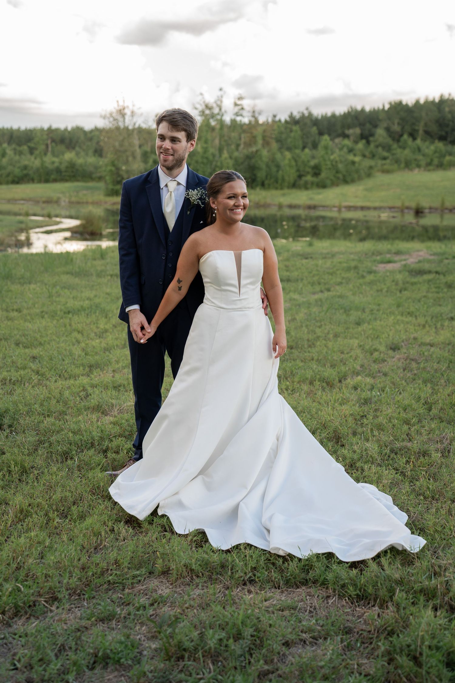 Bride in strapless white gown holds hands with groom in navy suit on lush green field at outdoor wedding.