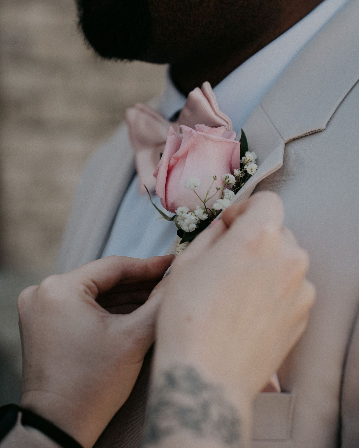 Hands pinning a pink rose boutonniere with baby's breath onto a gray suit lapel at a wedding.
