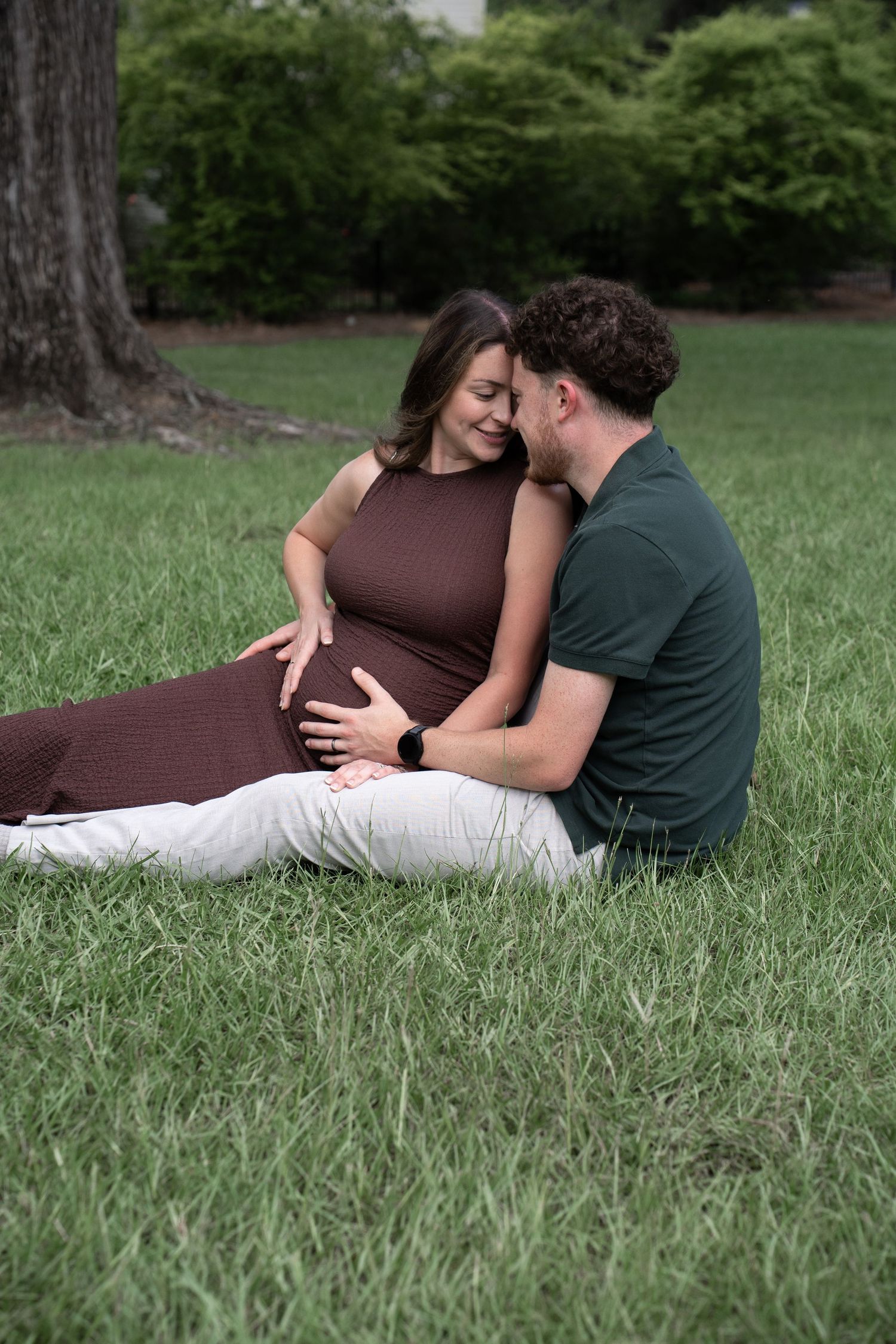 Pregnant woman in brown dress sits on grass with partner, both smiling and cradling her baby bump in a lush green park.