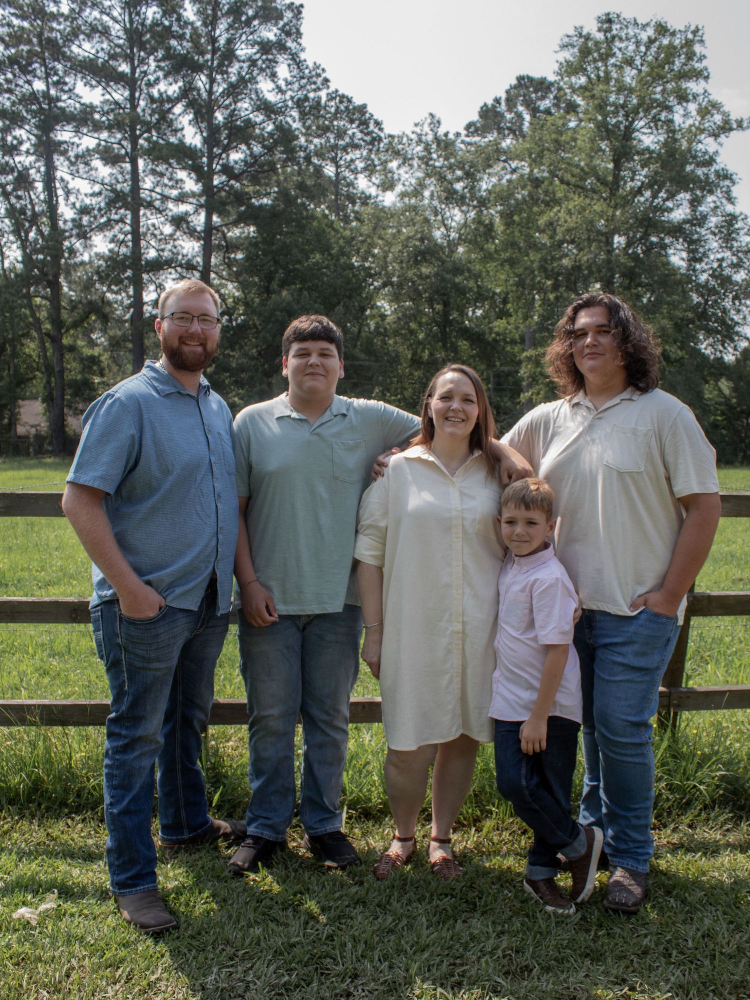 Family of five posing outdoors on green grass near wooden fence with tall trees in background on sunny day.