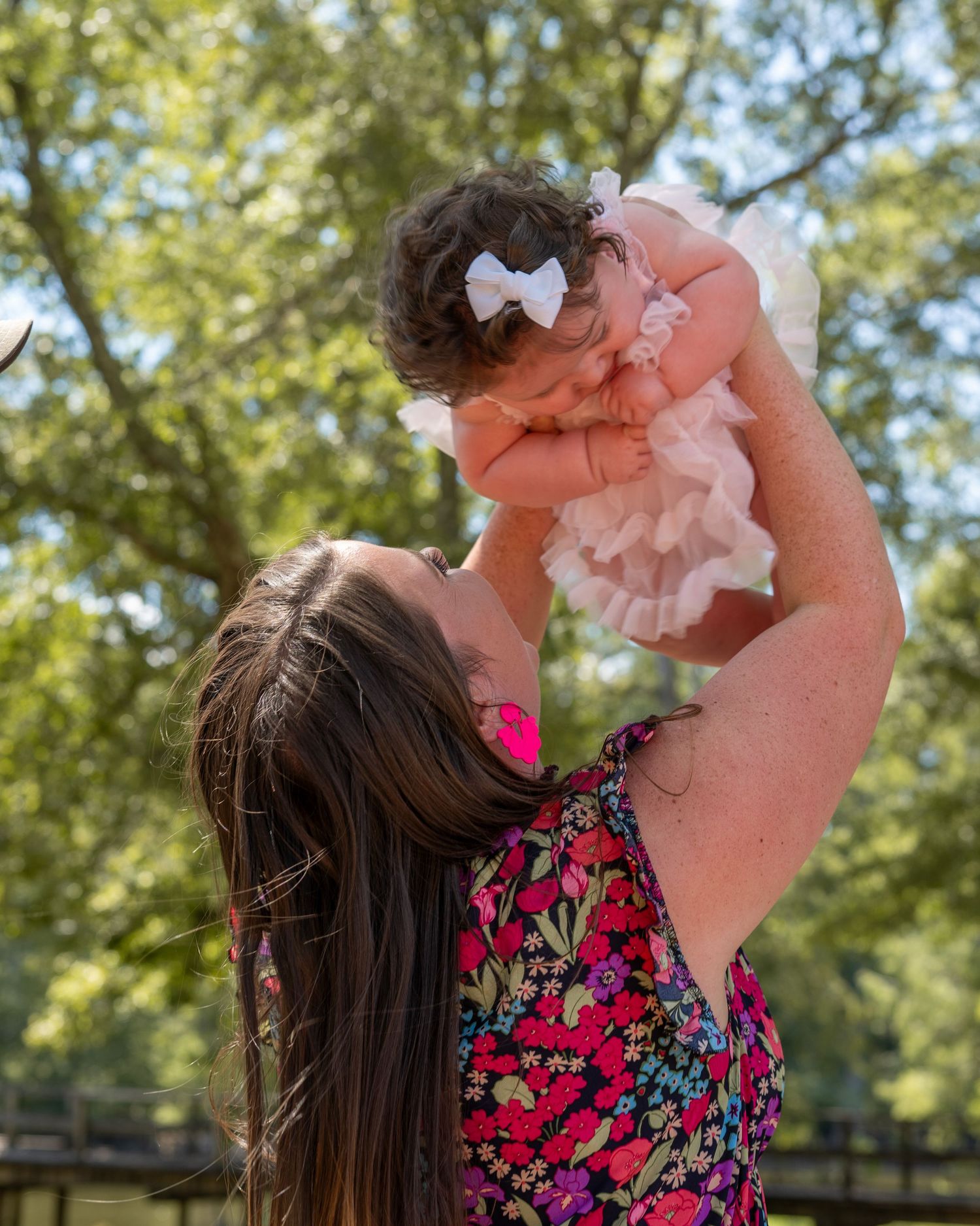 A mother joyfully lifts her baby girl in a white bow headband outdoors among lush green trees.