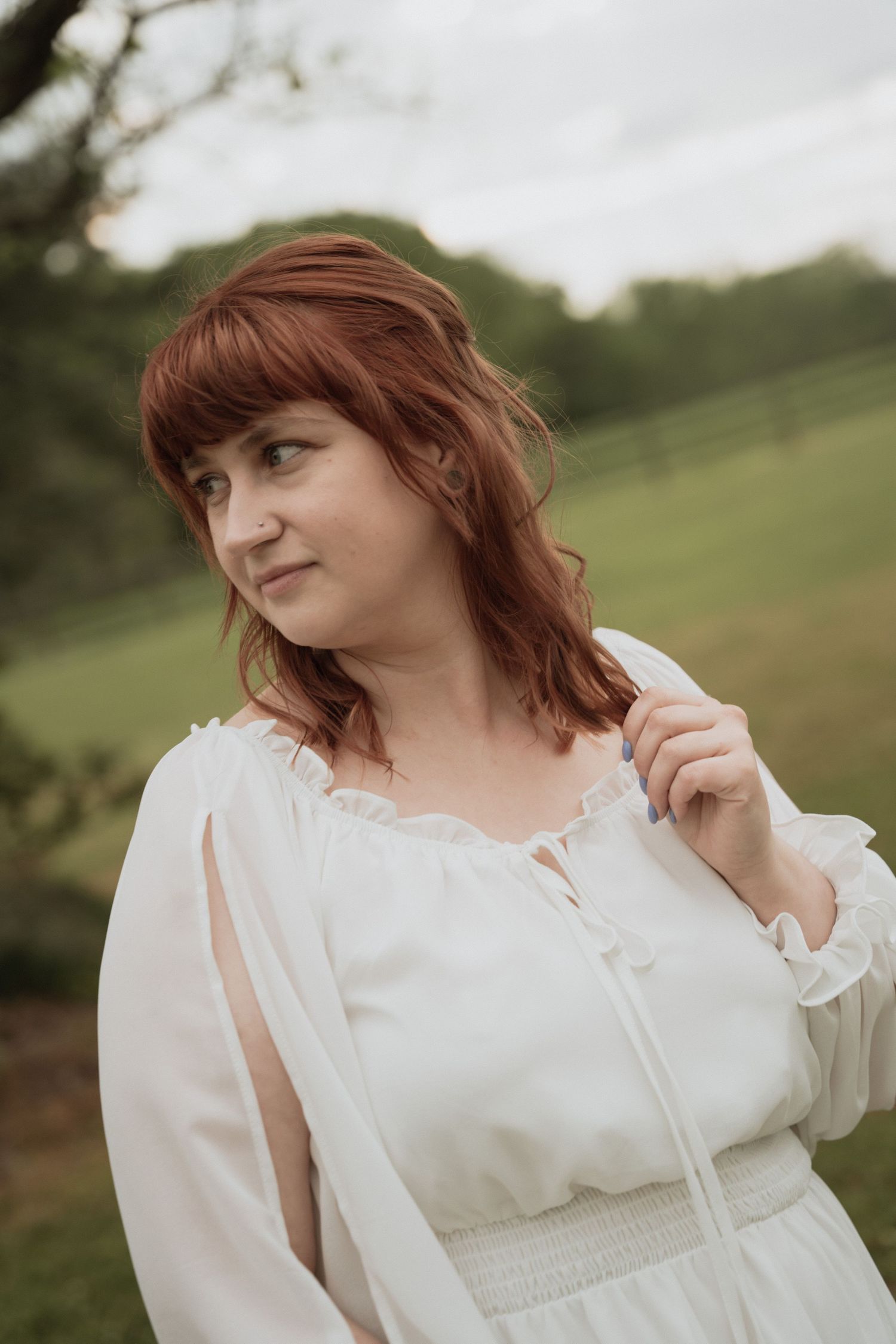 A young woman with red hair wearing a white boho dress stands outdoors in a green field, looking to the side.