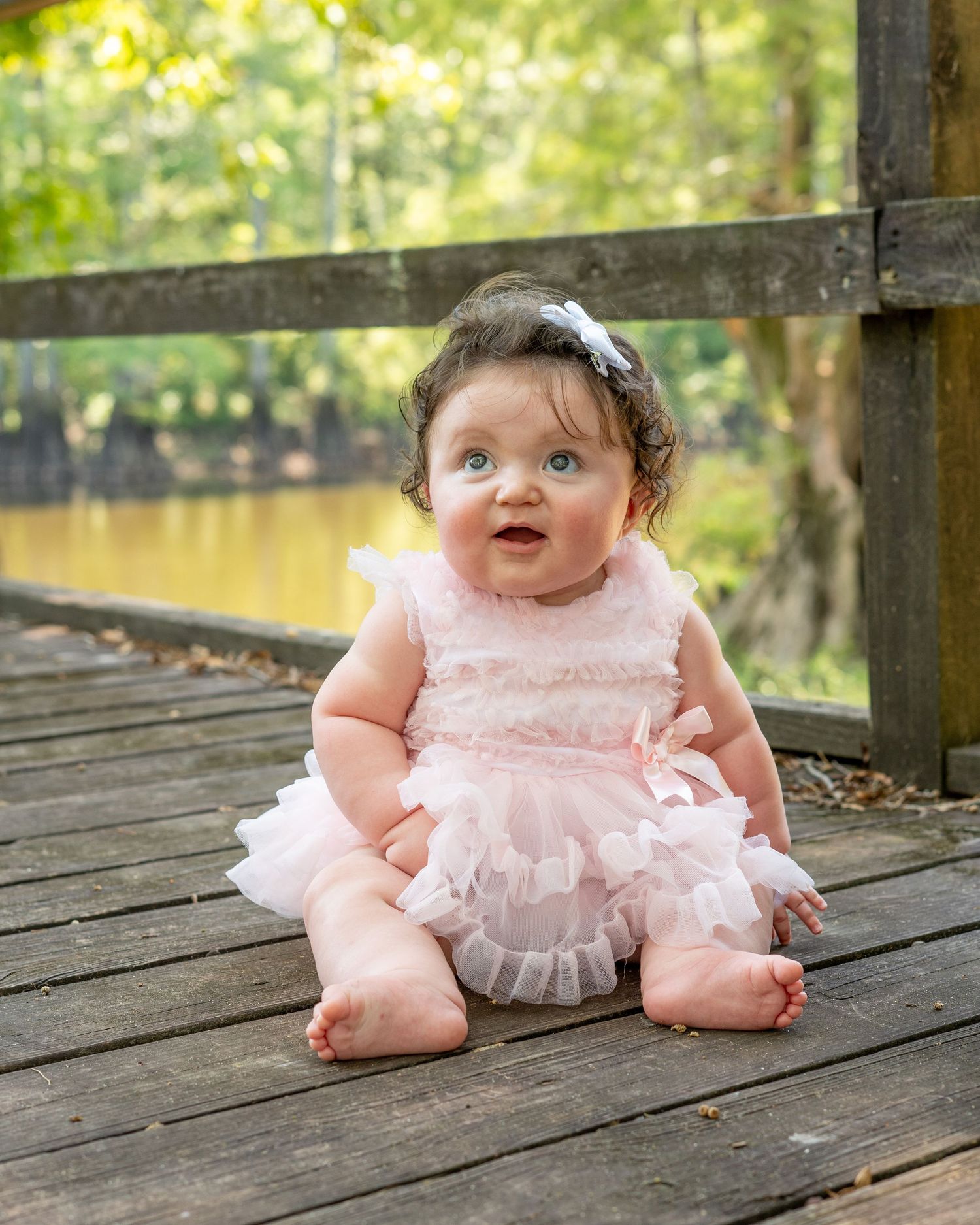 Adorable baby girl in pink tutu dress sitting on wooden deck outdoors, wearing white bow headband.