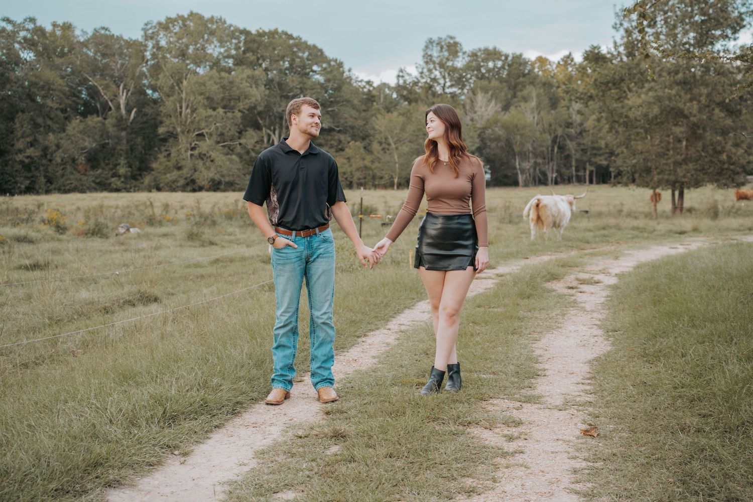 Couple holding hands walking on a dirt path in a green field with horses grazing in the background.