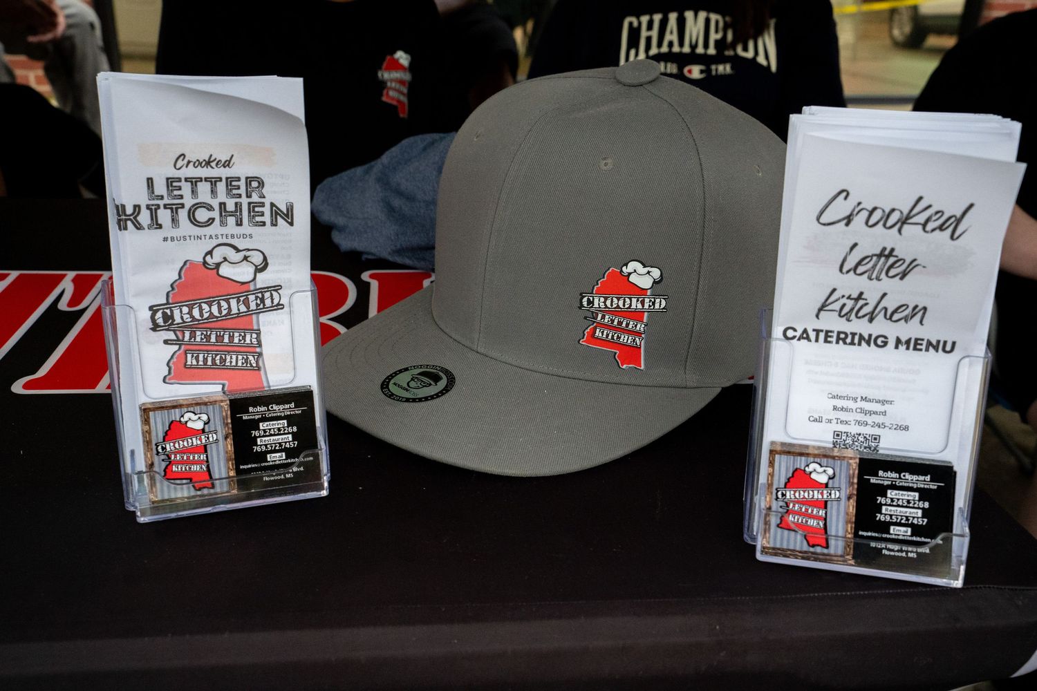 Crooked Letter Kitchen catering menu display with branded gray snapback hat and promotional materials on a table.