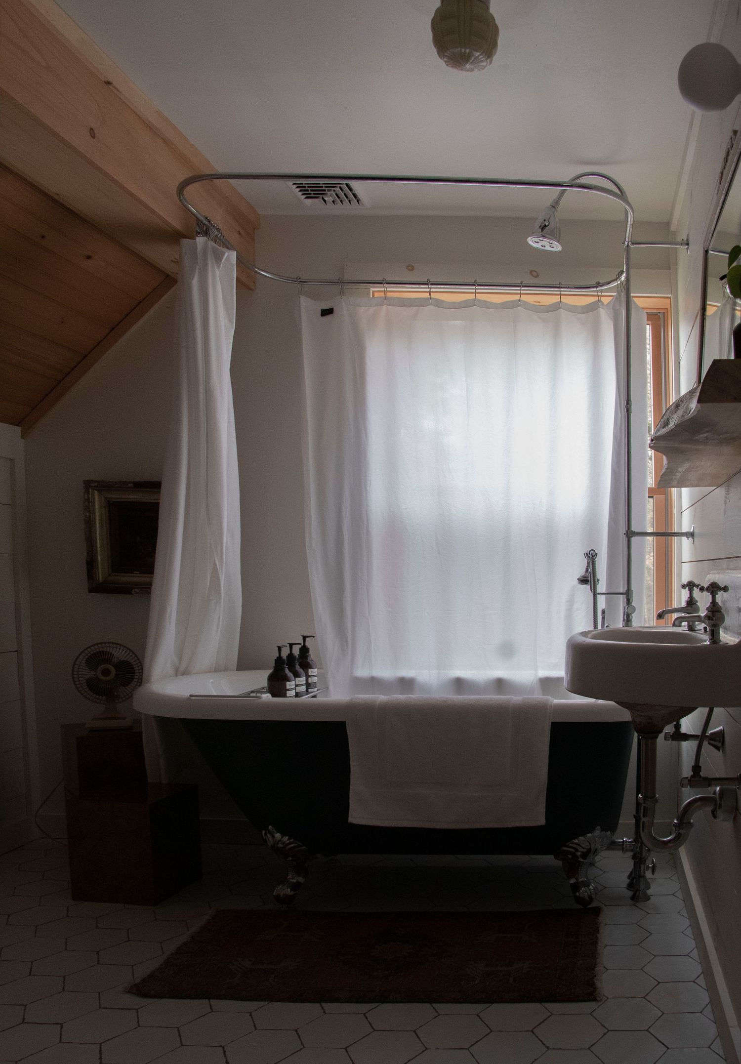 Vintage clawfoot bathtub with white curtains, wood ceiling, and pedestal sink in a rustic bathroom.