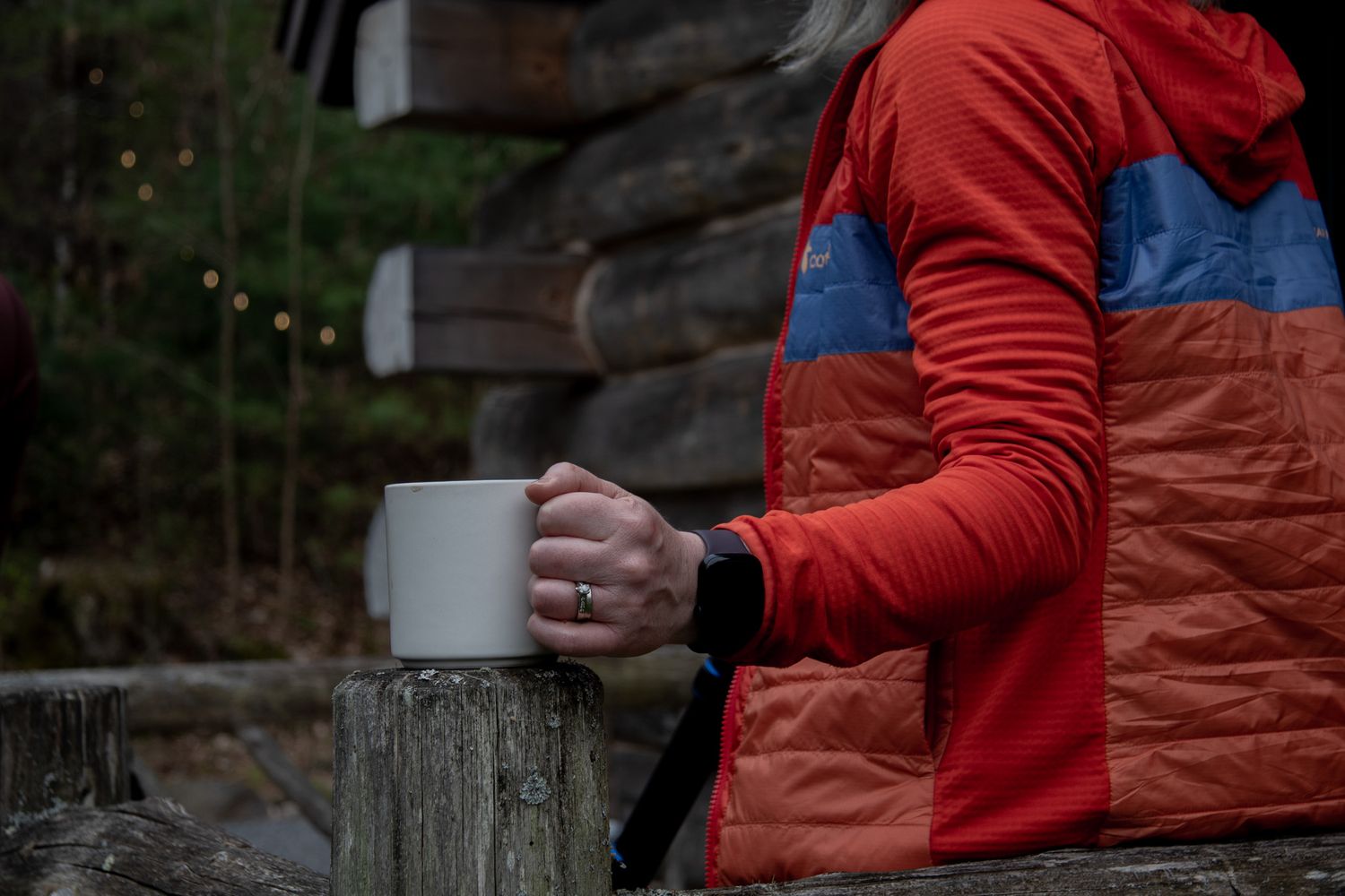 Person in red jacket holding a white mug outdoors near a rustic log cabin in a forest setting.