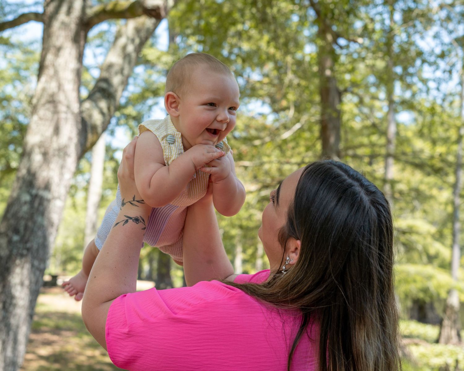 A smiling baby being lifted up by a woman in a pink shirt outdoors in a sunny tree-filled park.