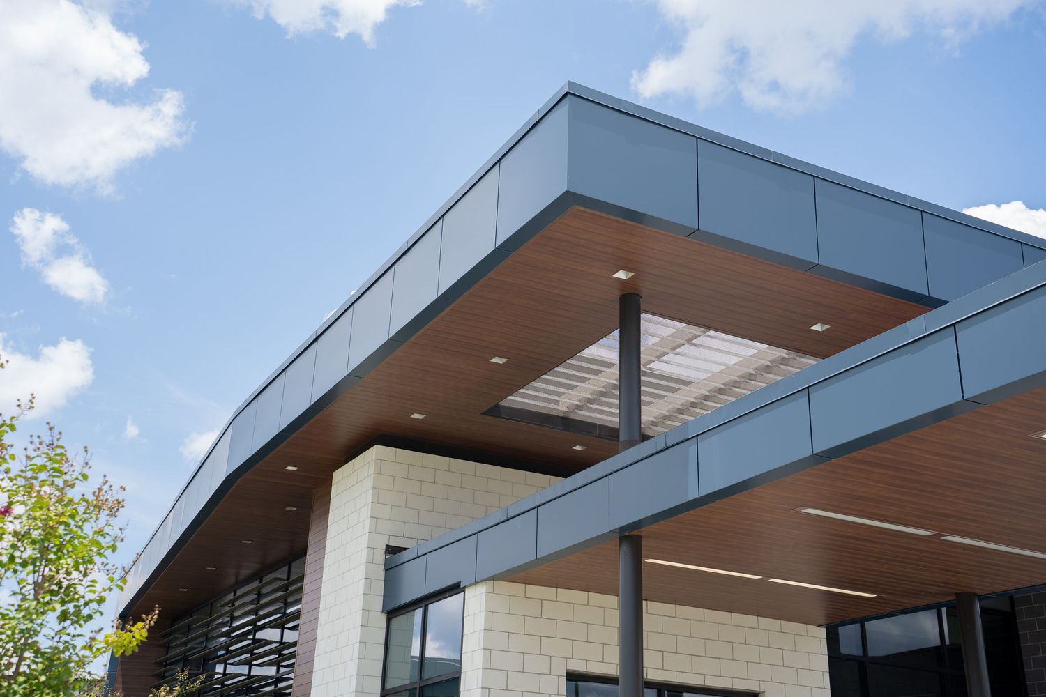 Modern commercial building exterior featuring wood ceiling panels, gray metal facade, and large windows under a blue sky.
