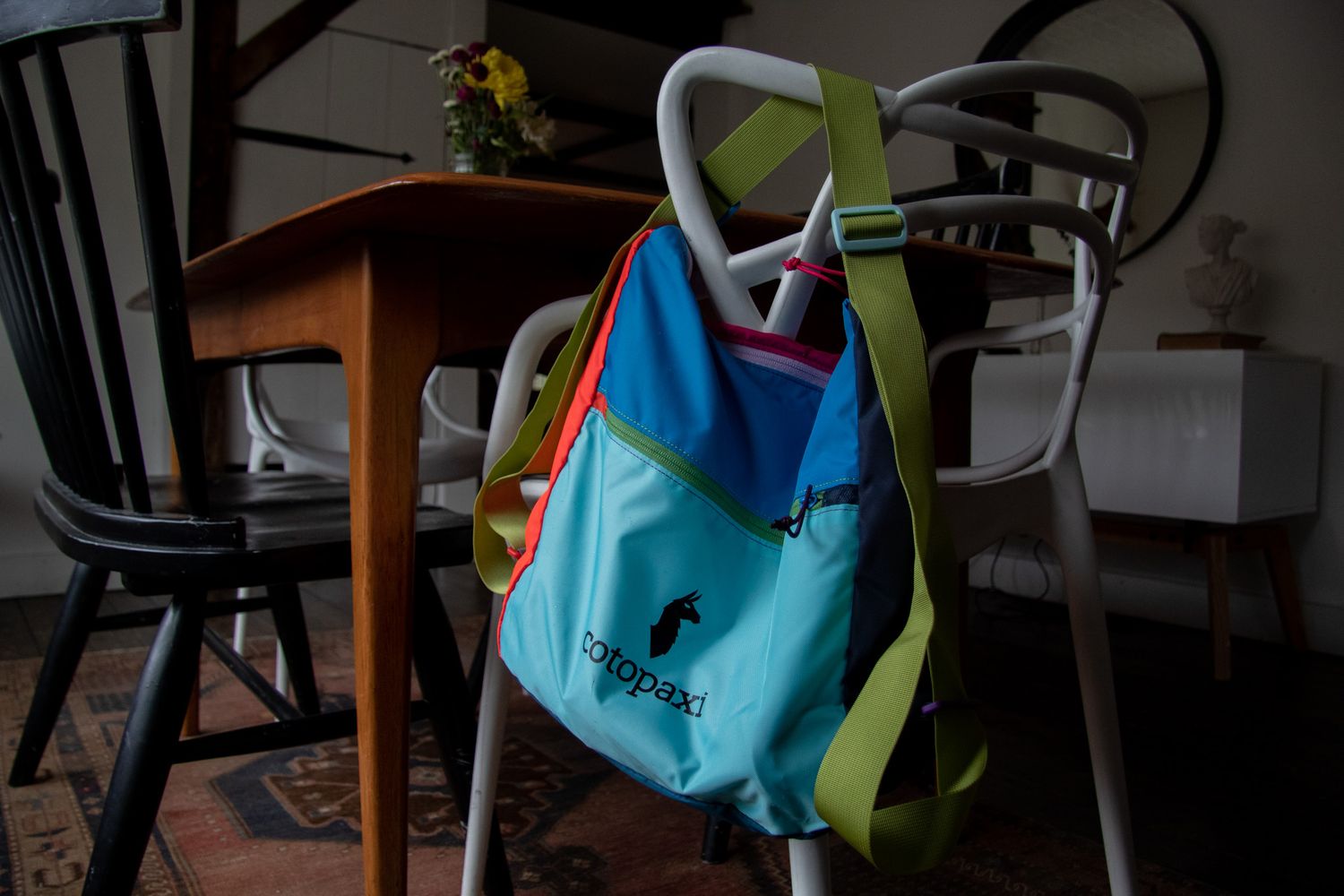 Colorful Cotopaxi tote bag hanging on a white chair in a cozy dining room with wooden table.
