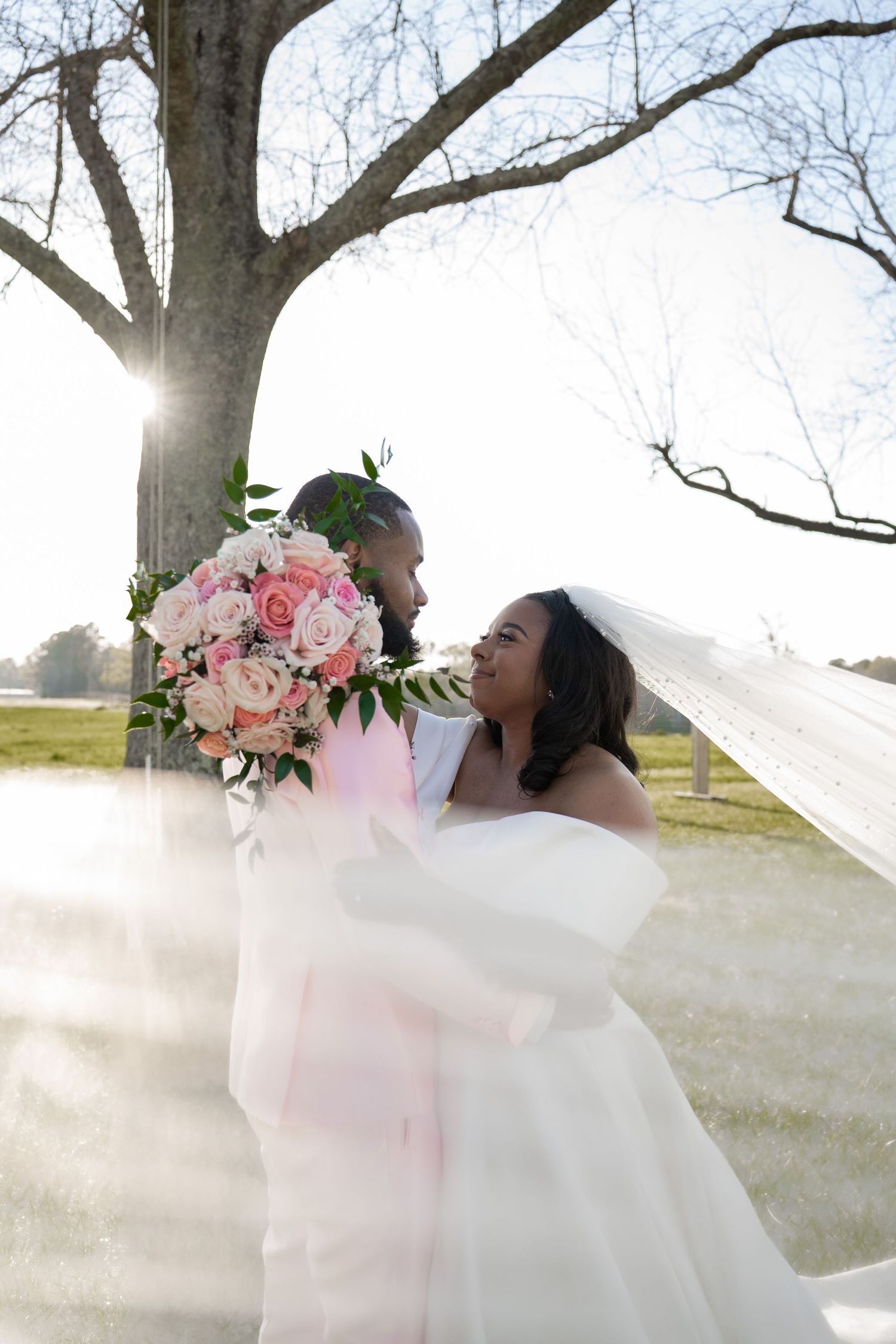 A joyful Black bride and groom share a romantic moment outdoors, with her pink rose bouquet and flowing veil catching sunlight.