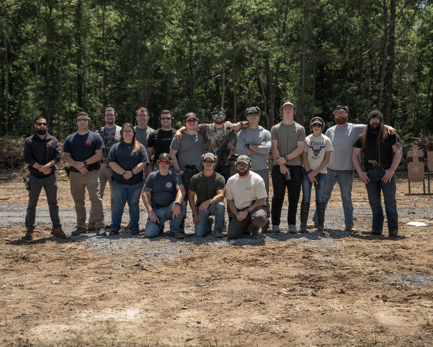 Group of people posing together outdoors at a shooting range surrounded by trees on a sunny day.
