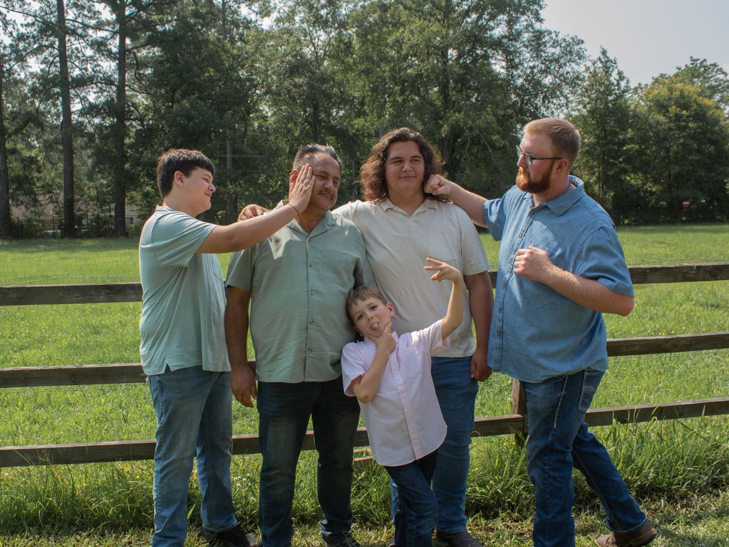 Family of five posing outdoors near a wooden fence on a green farm, wearing casual blue and white shirts.