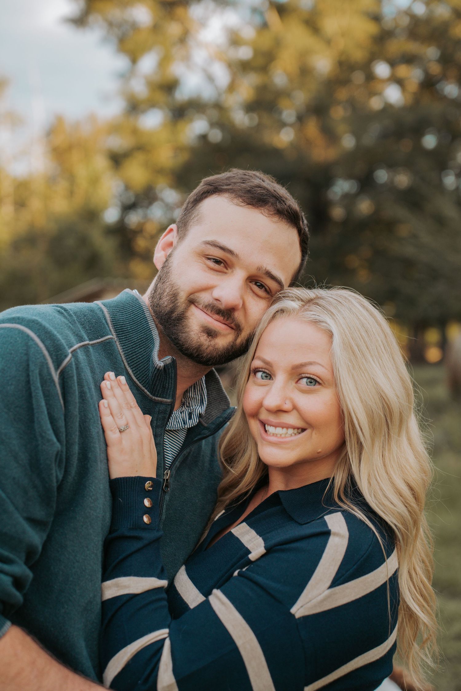 A smiling couple poses outdoors in fall, woman in striped sweater, man in teal pullover, surrounded by autumn foliage.