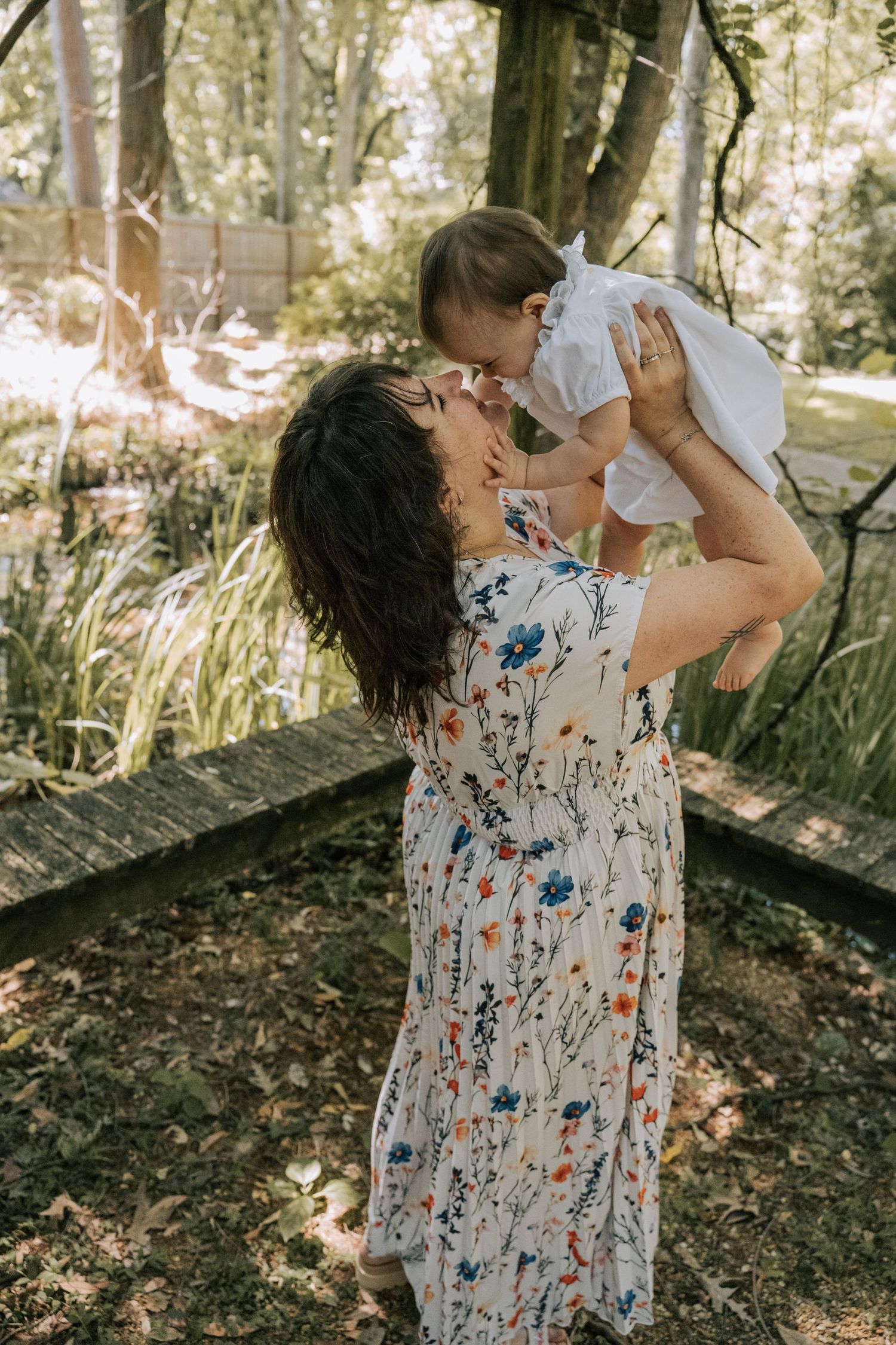 A mother in a floral dress lifts her baby up for a kiss outdoors in a lush green garden setting.