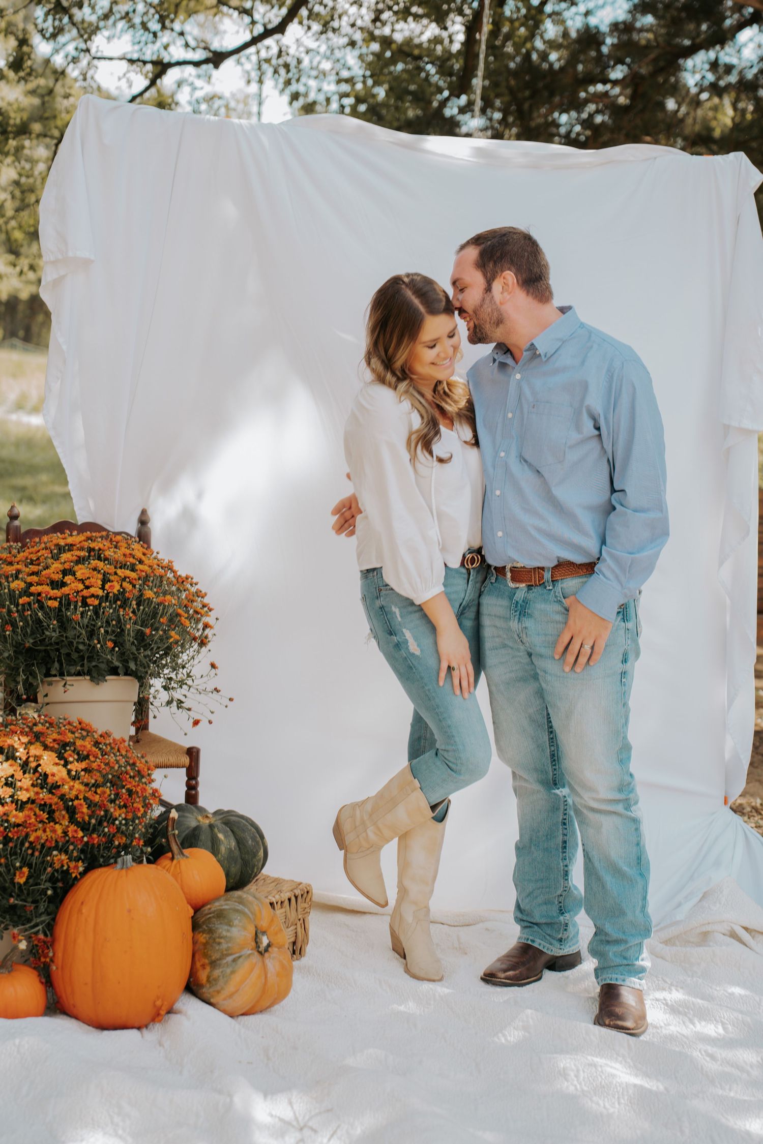 Couple in jeans poses romantically by white backdrop with fall pumpkins and orange mums in outdoor autumn photo session.