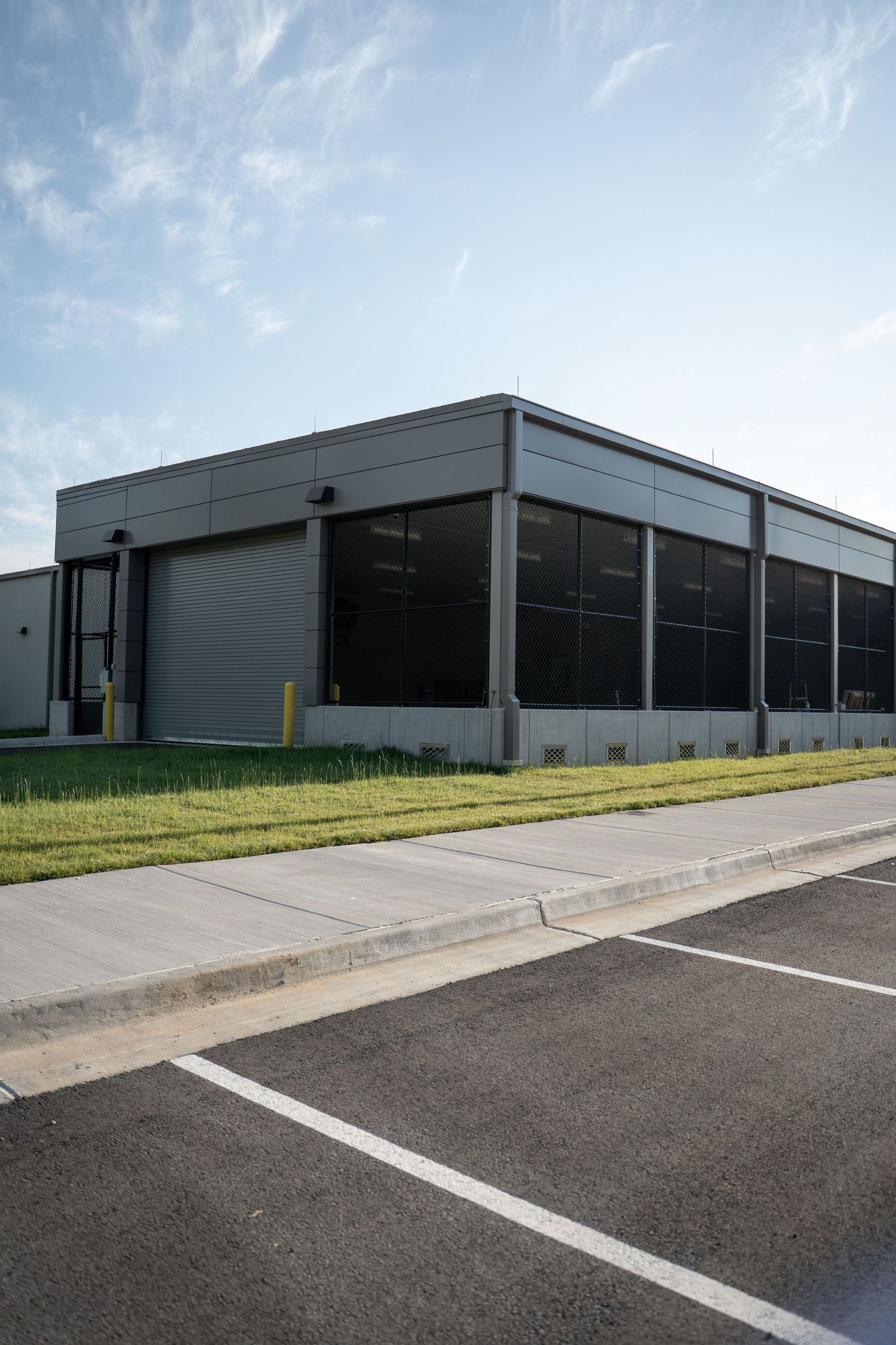 Modern industrial warehouse building with large windows, parking lot, and manicured lawn under a partly cloudy sky.