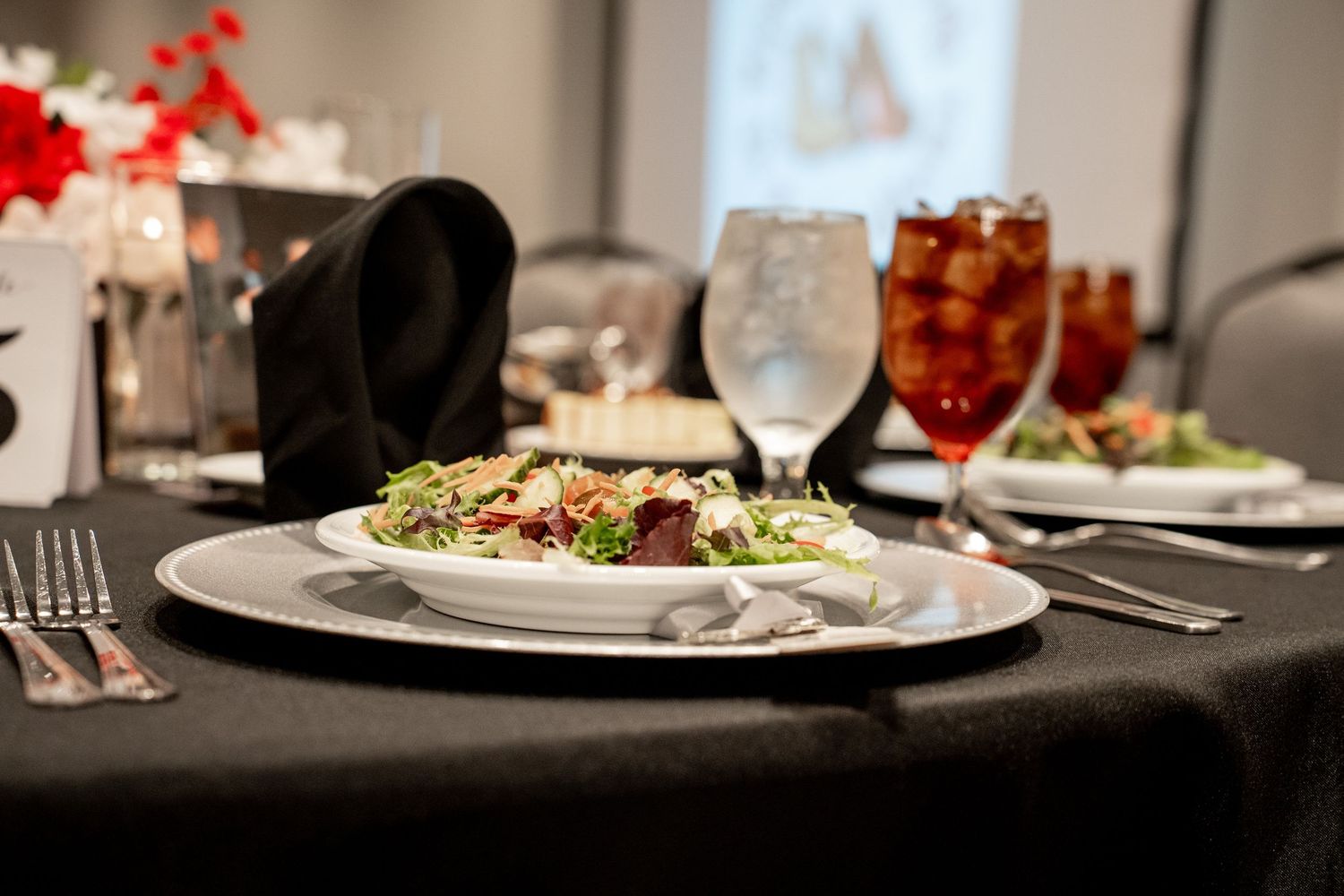 Elegant wedding reception place setting with fresh salad, glassware, and black napkin on a dark tablecloth.