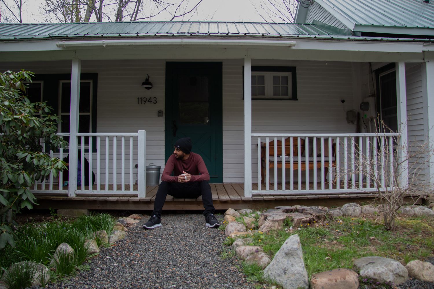 Man sitting alone on front porch steps of white house with teal door, appearing contemplative on overcast day.