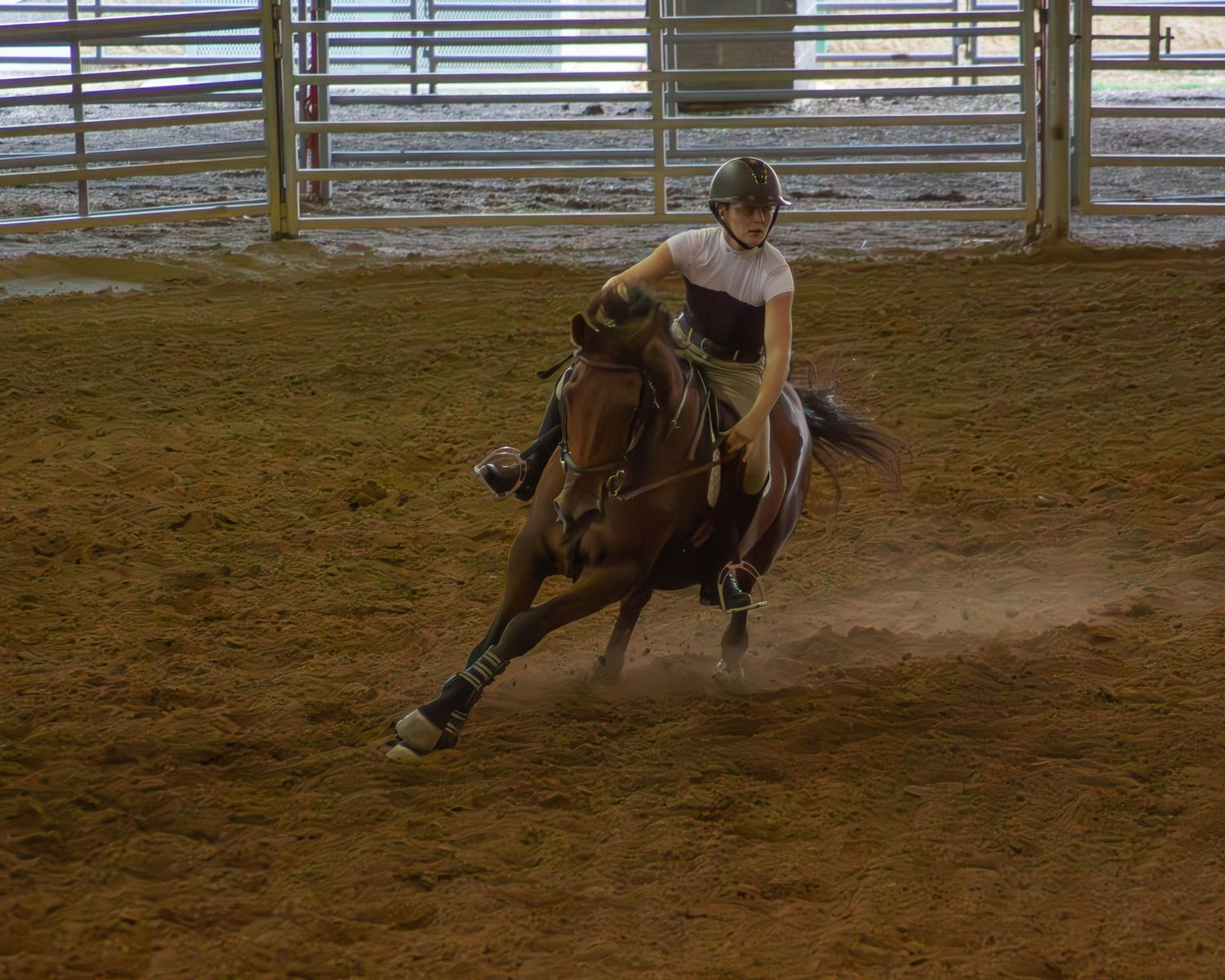 A rider in a helmet competes in barrel racing on a brown horse in an indoor dirt arena.