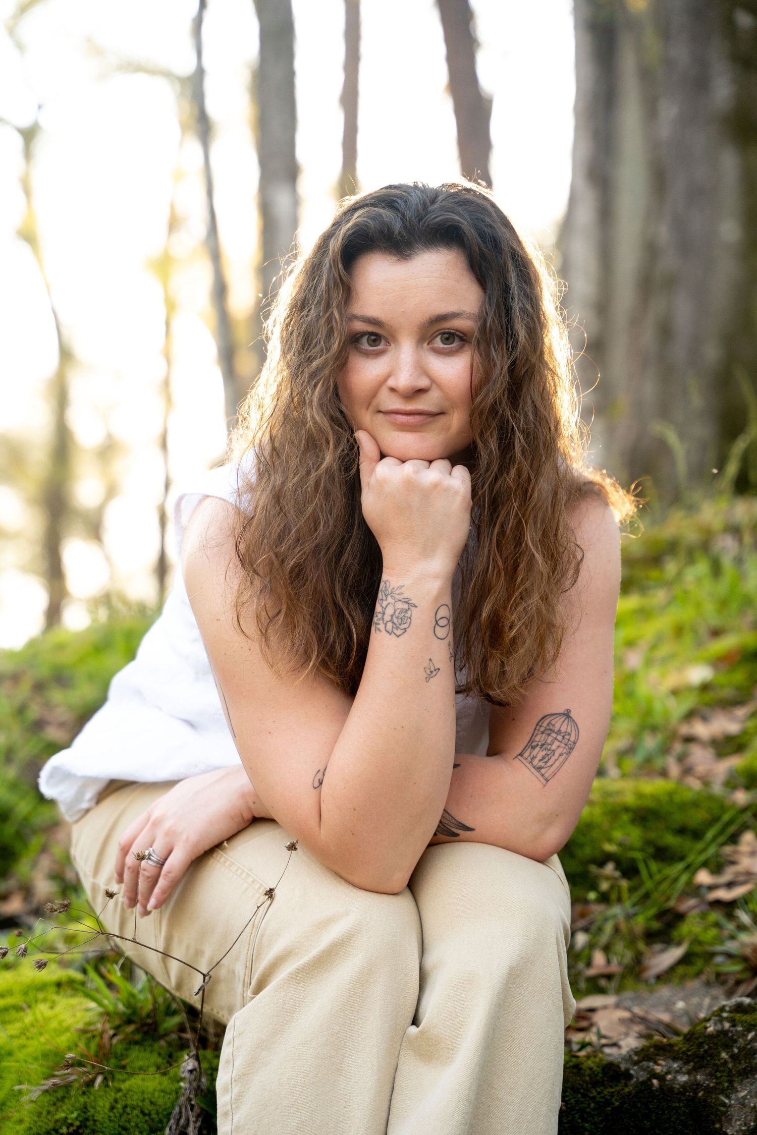 Woman with curly brown hair and tattoos sits on mossy forest ground, resting chin on hand.
