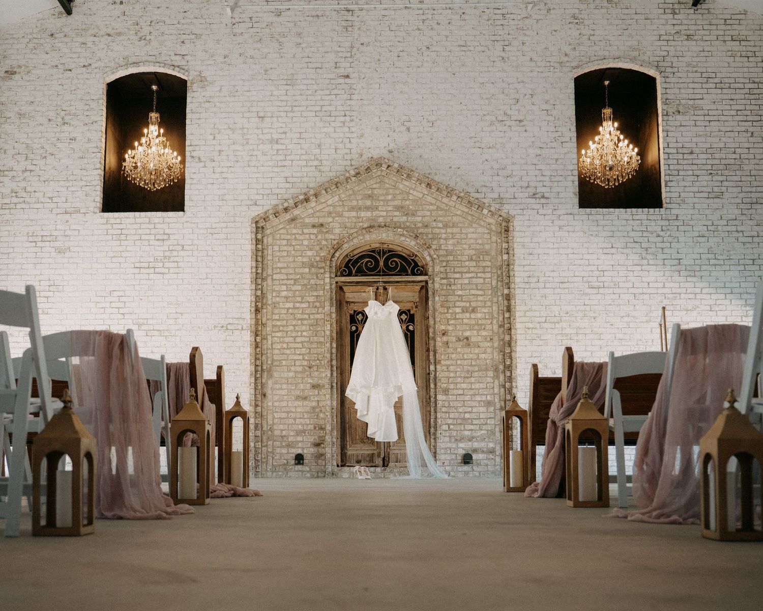 Wedding dress hanging in a rustic white brick chapel with wooden pews, draped fabric, and elegant chandeliers.
