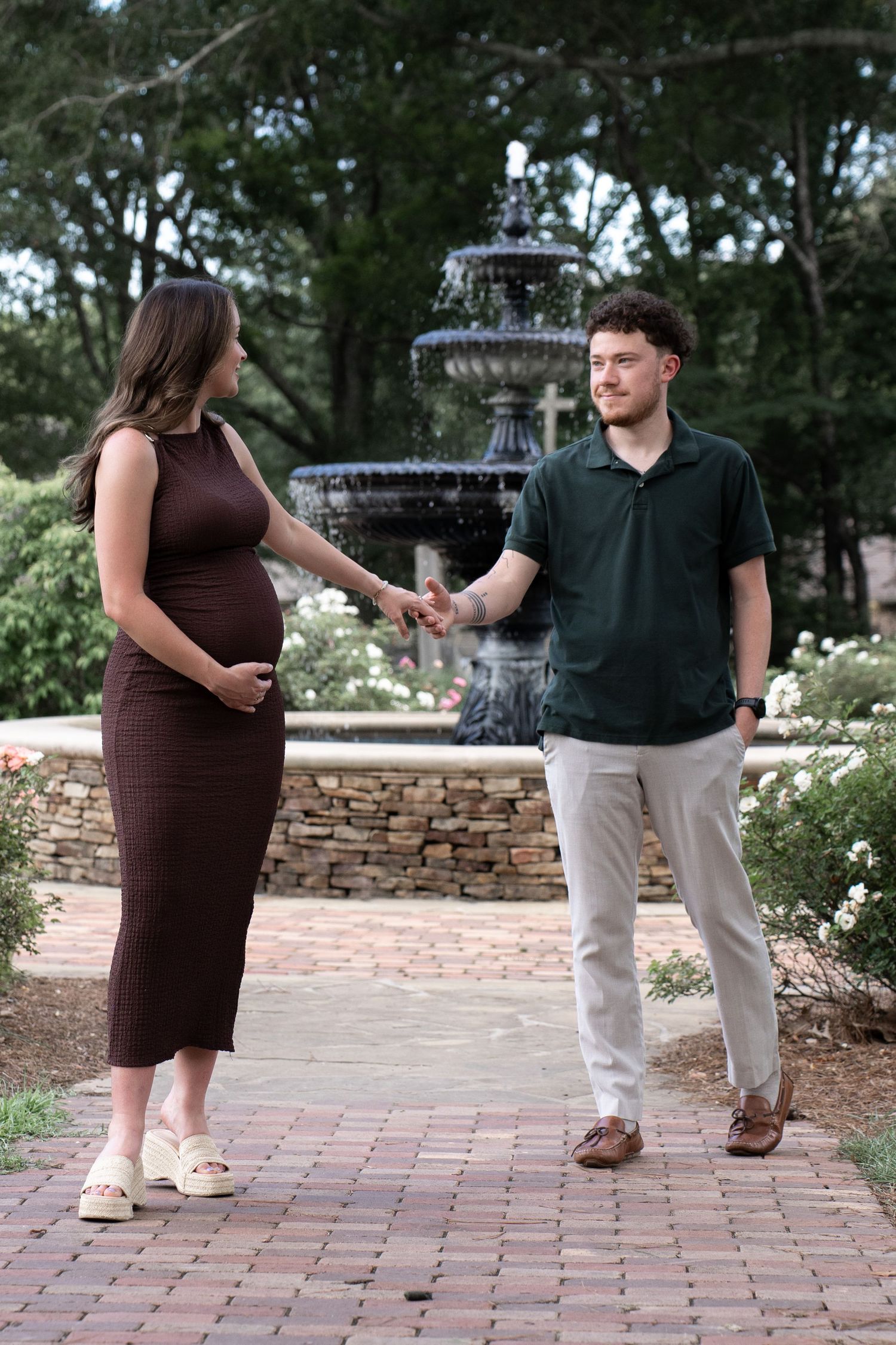 Pregnant woman in brown dress holds hands with man in green polo near stone fountain in garden.