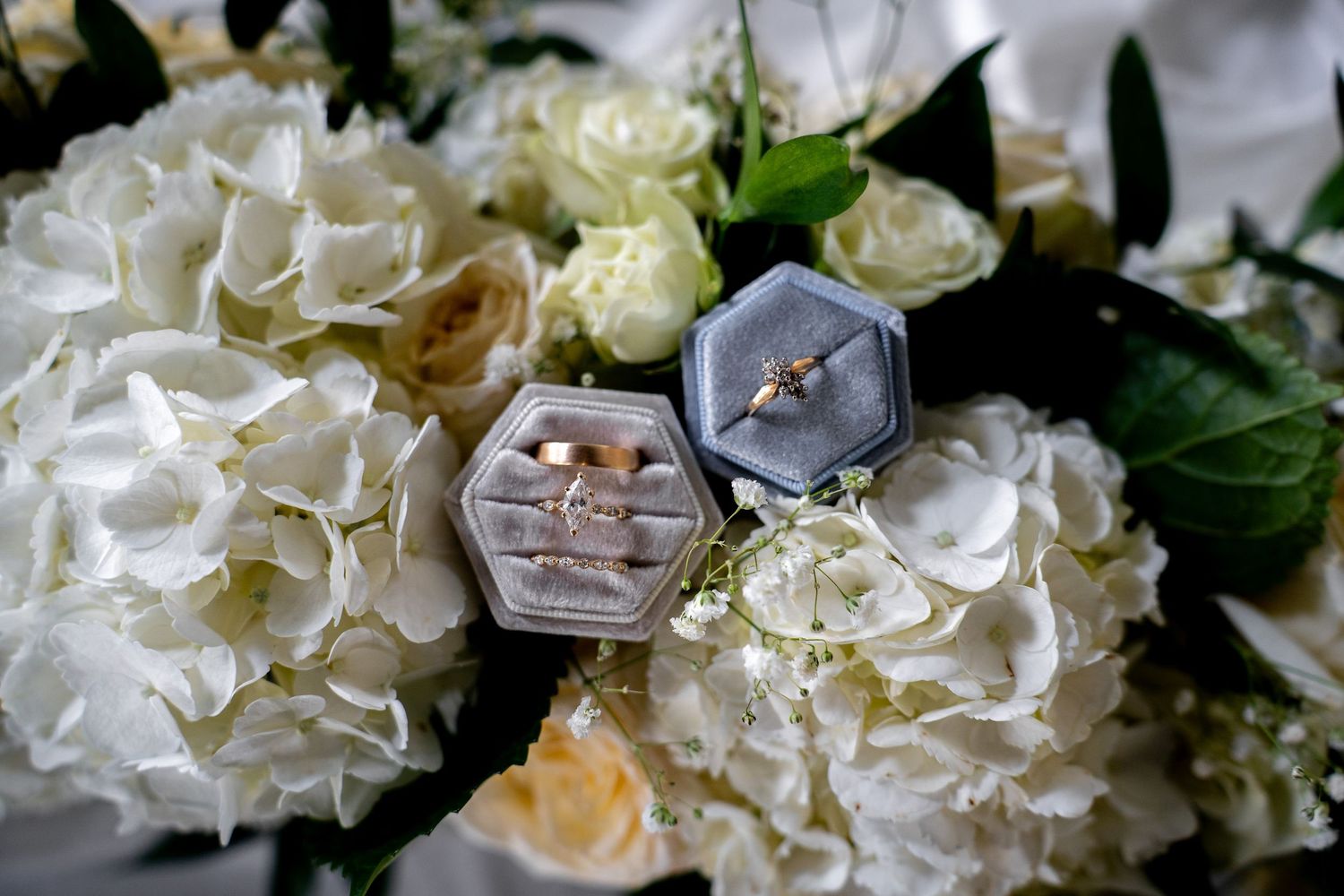 Wedding rings displayed in a gray velvet hexagon box atop a white hydrangea and rose bridal bouquet.